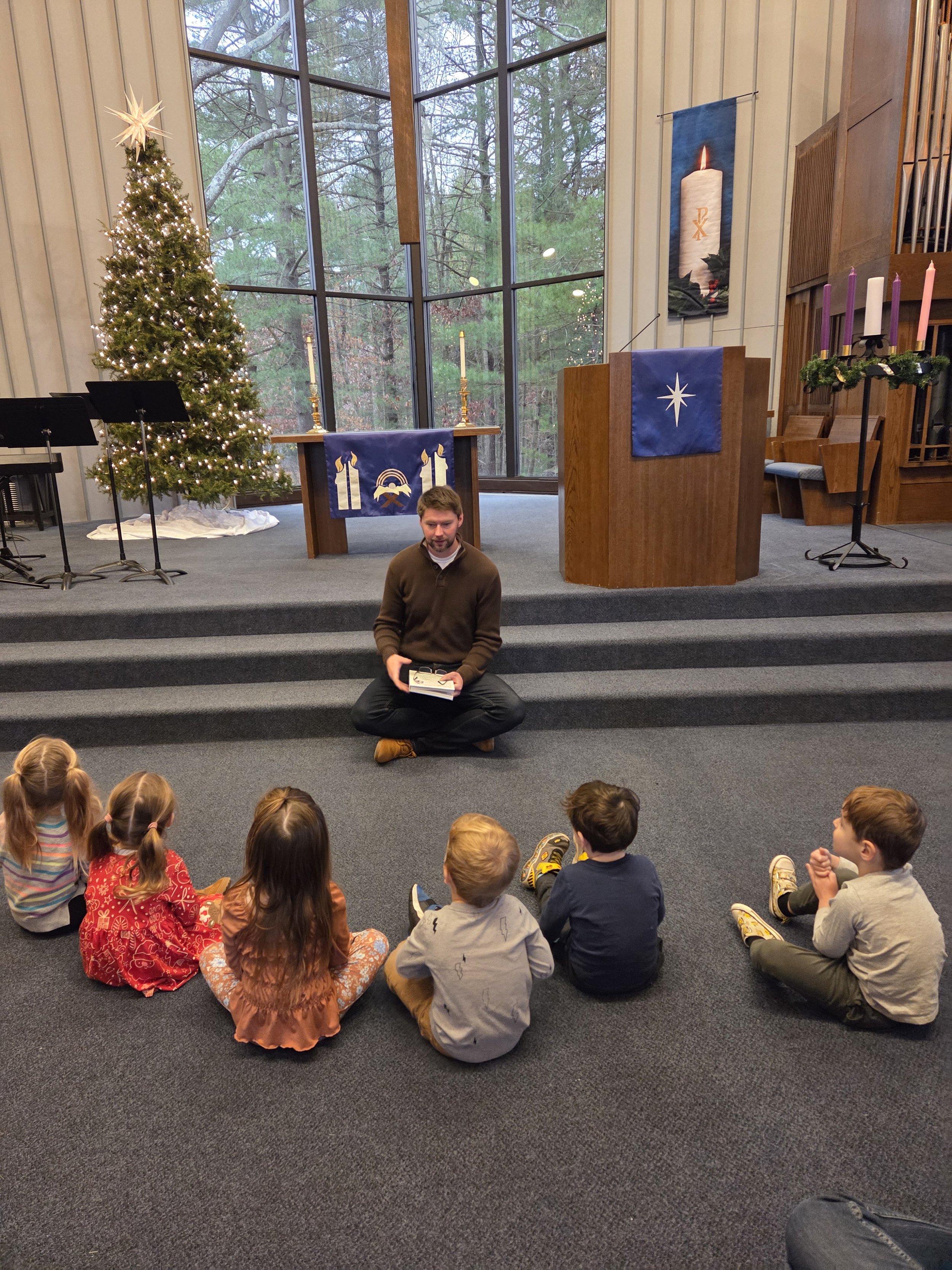 Inside a church decorated for Christmas with a Christmas tree, candles, and banners, a pastor sits on the stairs with children sitting on the floor in front of him.