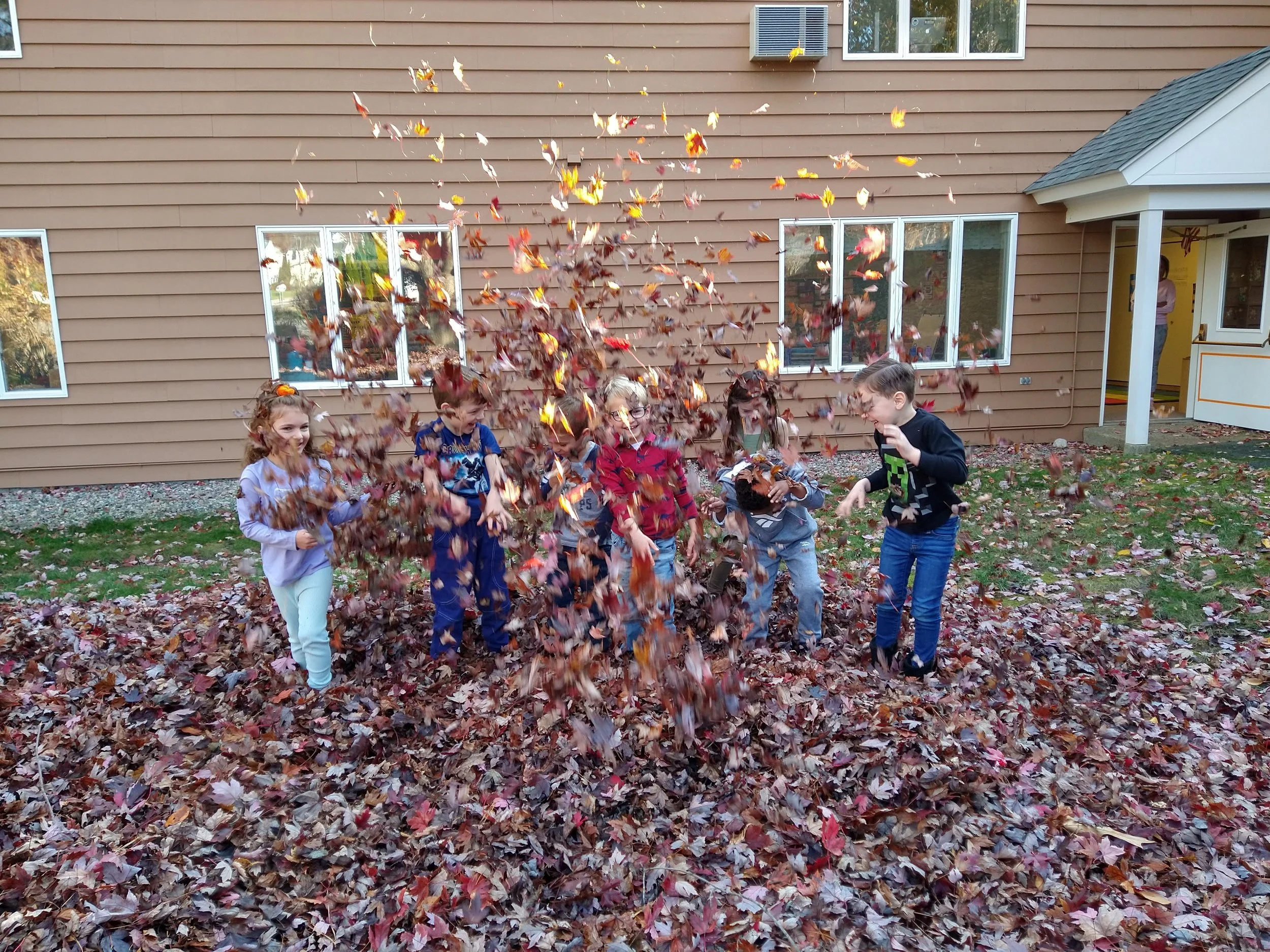 Children playing in a pile of fallen autumn leaves in a yard with a building wall, windows, and an air conditioning unit visible in the background.
