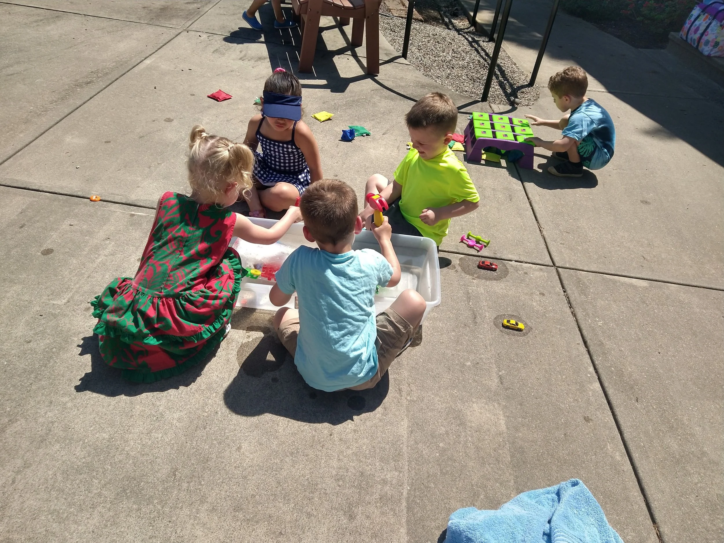 Children playing a game outdoors on a concrete pavement, with scattered toys and blankets, during a sunny day.