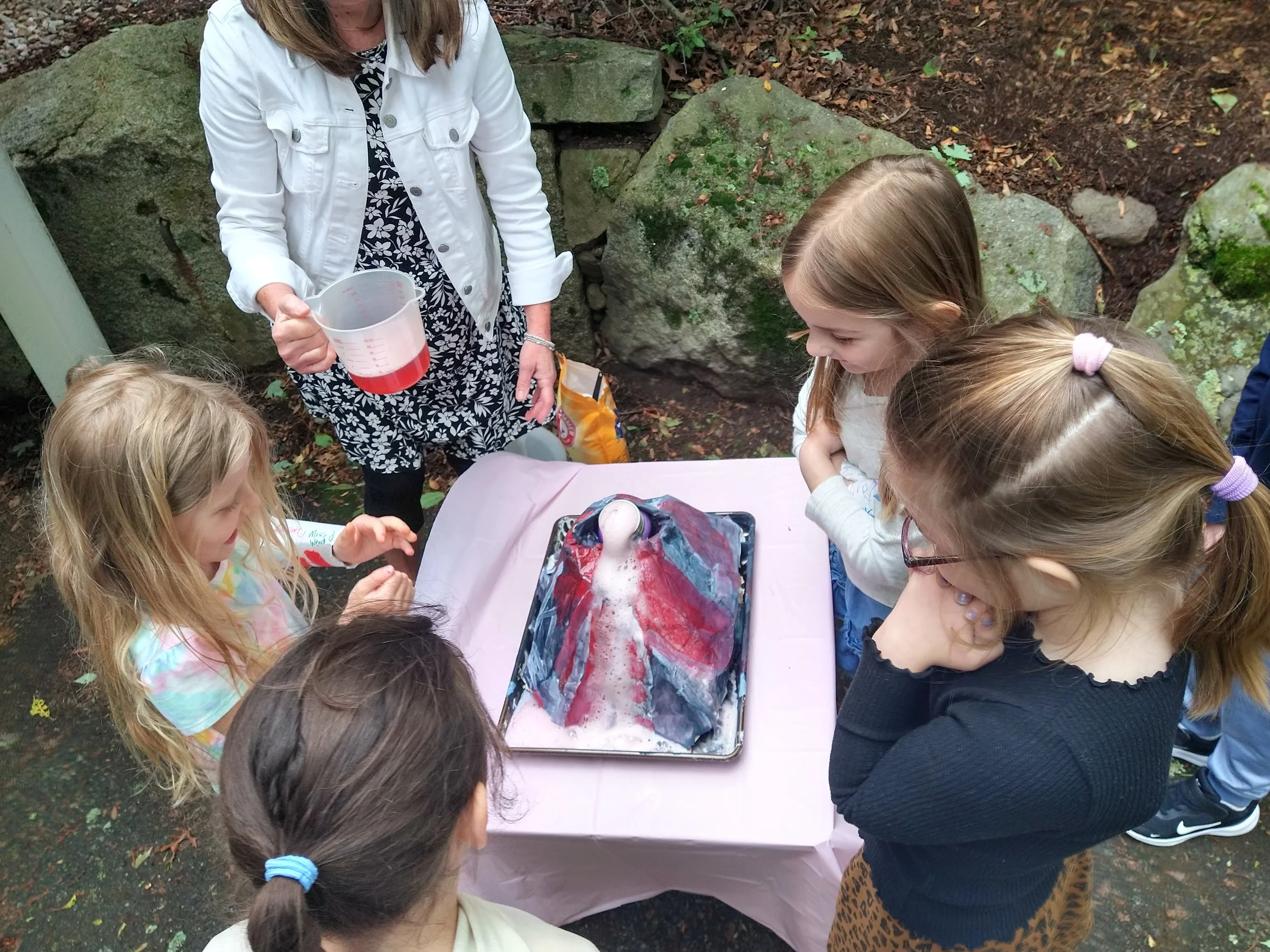 A group of children gathered around a table outdoors, observing a homemade craft volcano, with an adult woman pouring a red liquid into a clear plastic measuring cup.