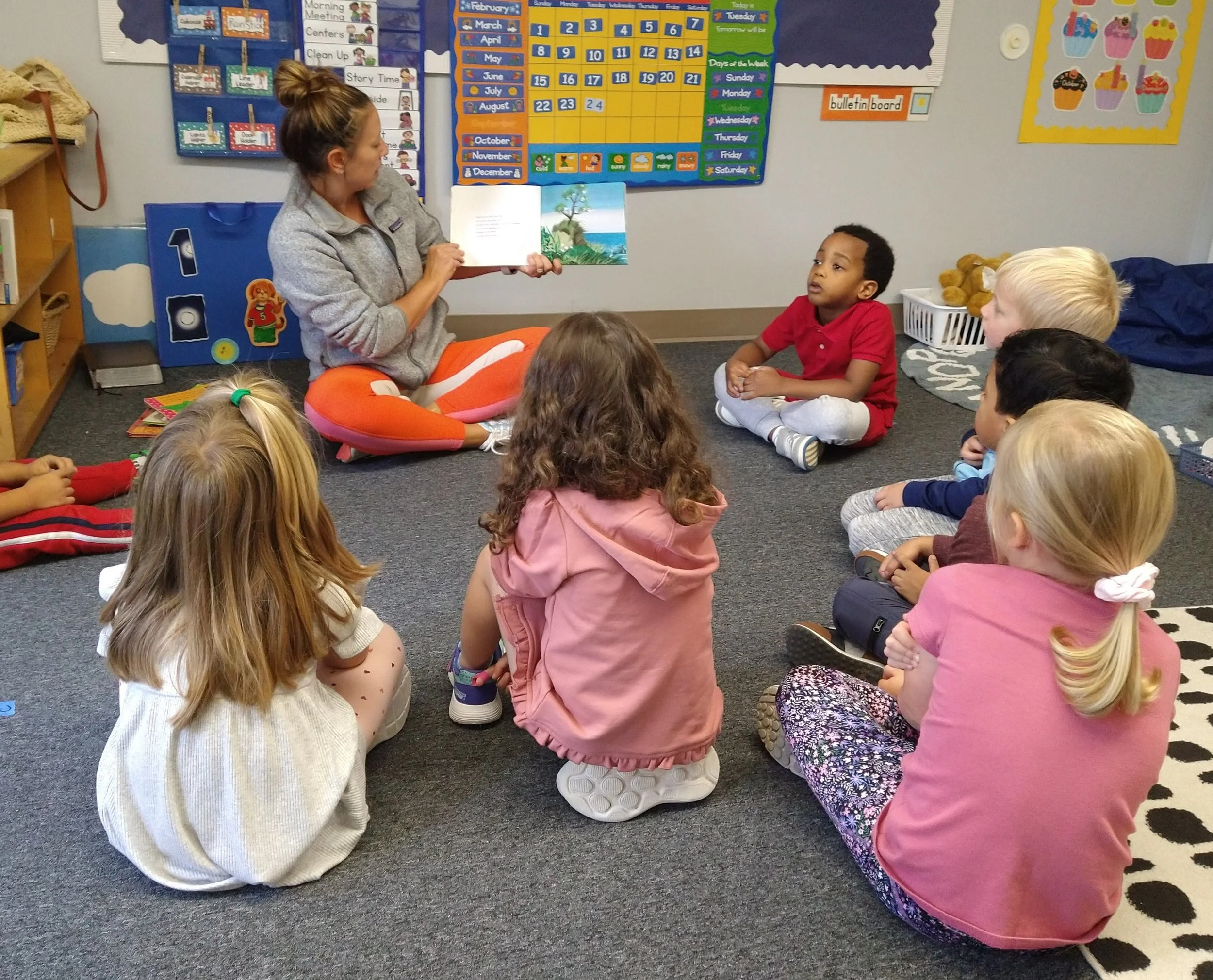 A woman reading a picture book to a group of young children in a classroom with colorful educational posters and a calendar on the wall.