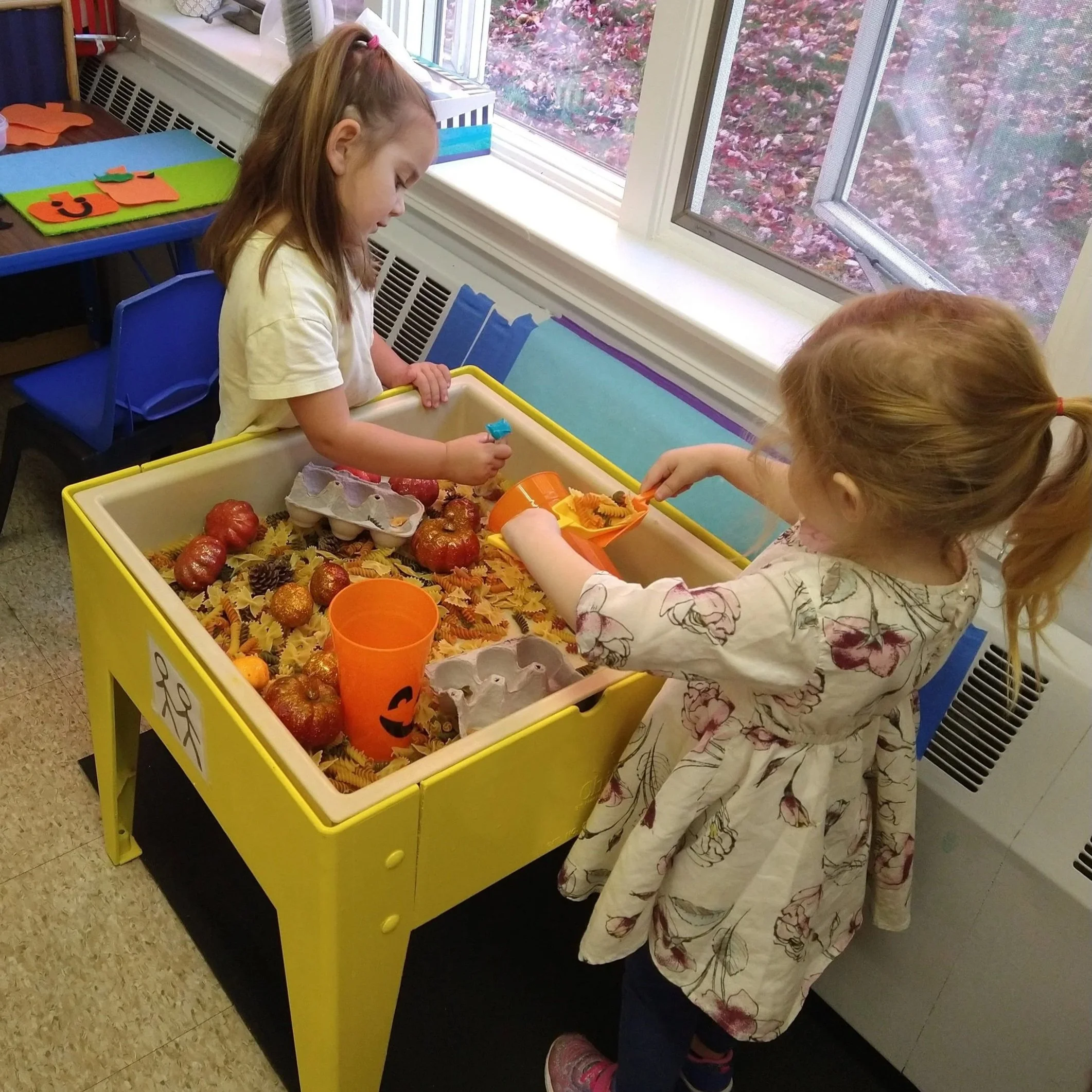 Children sorting Halloween pumpkins and decorations in a classroom with a large window showing autumn leaves outside.