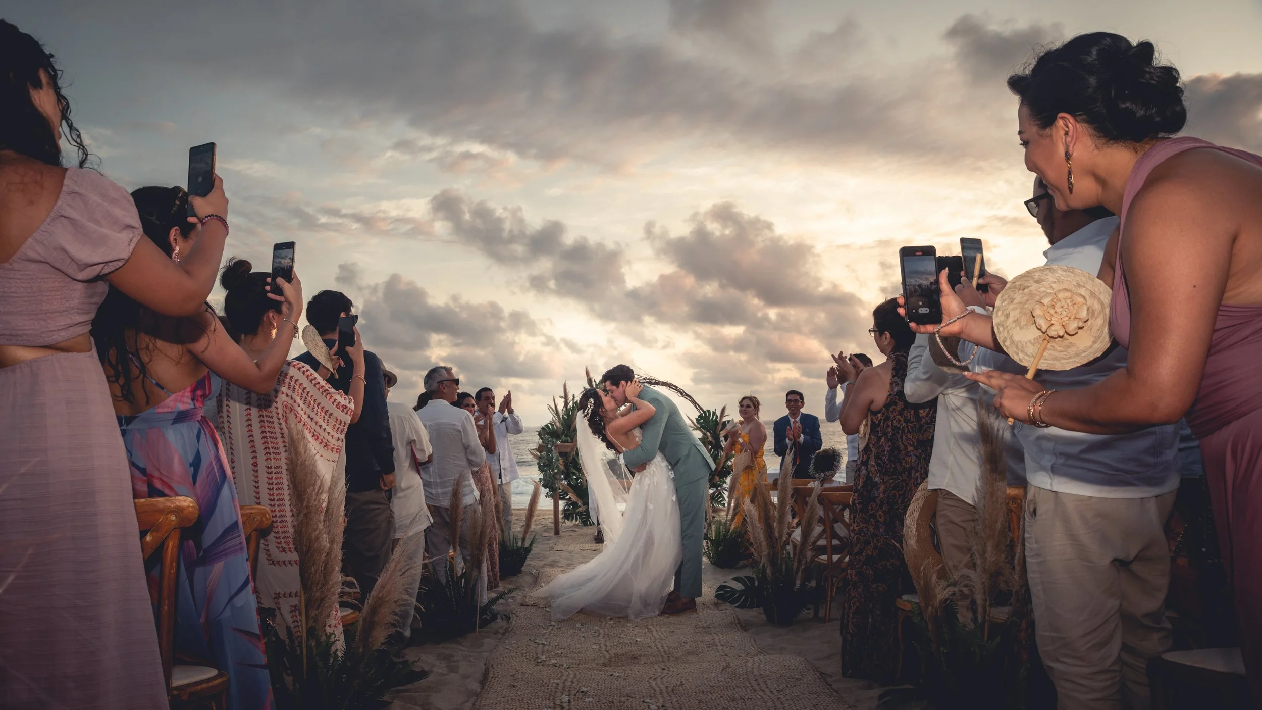 End of the ceremony of a  wedding in Puerto Escondido where the couple are embracing and kissing in front of their guests all around them in a beautiful sunset in front of the ocean in Puerto Escondido.