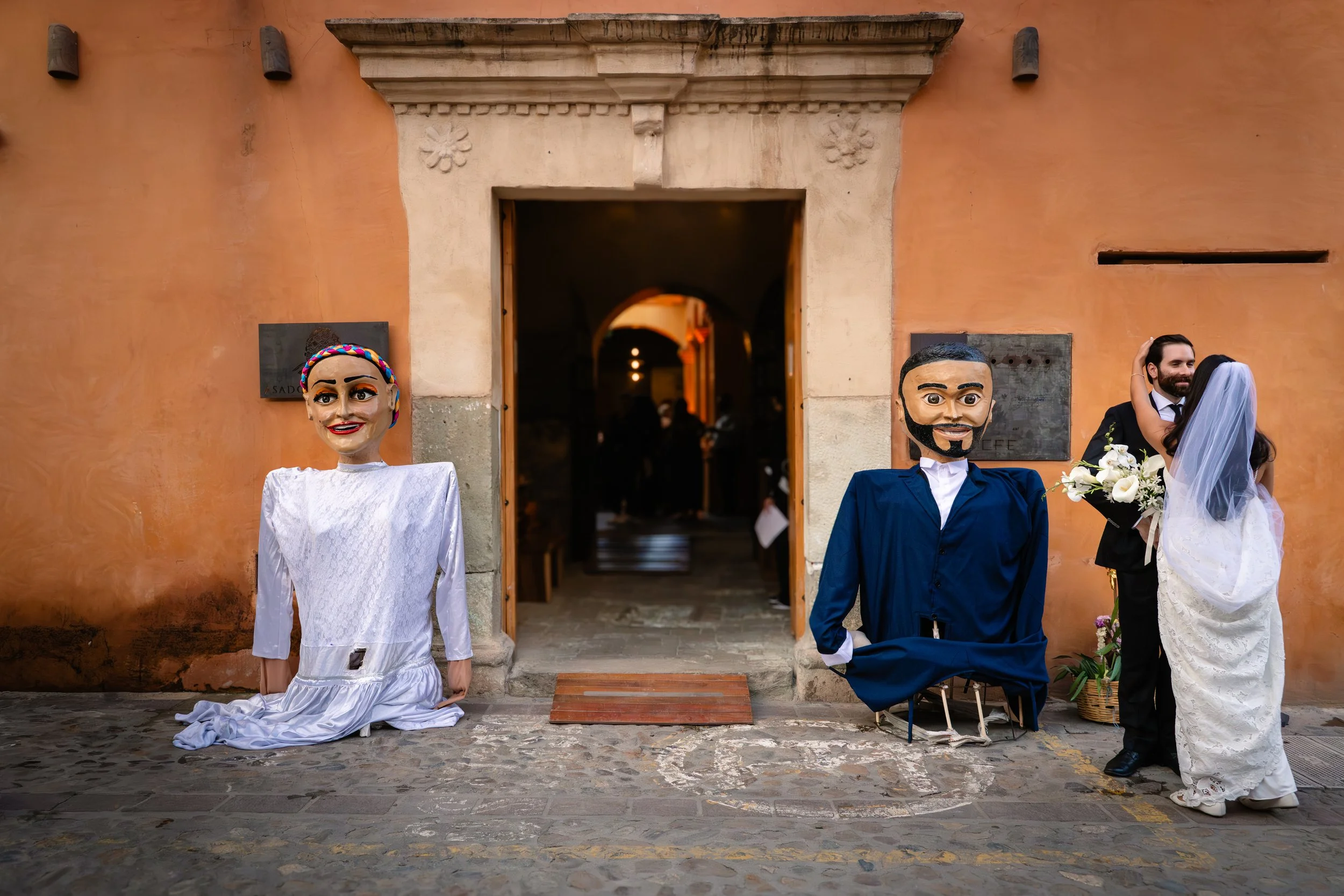 Maried couple in front of restaurant with their clalenda traditional puppet for their wedding. Oaxacan luxury wedding photography