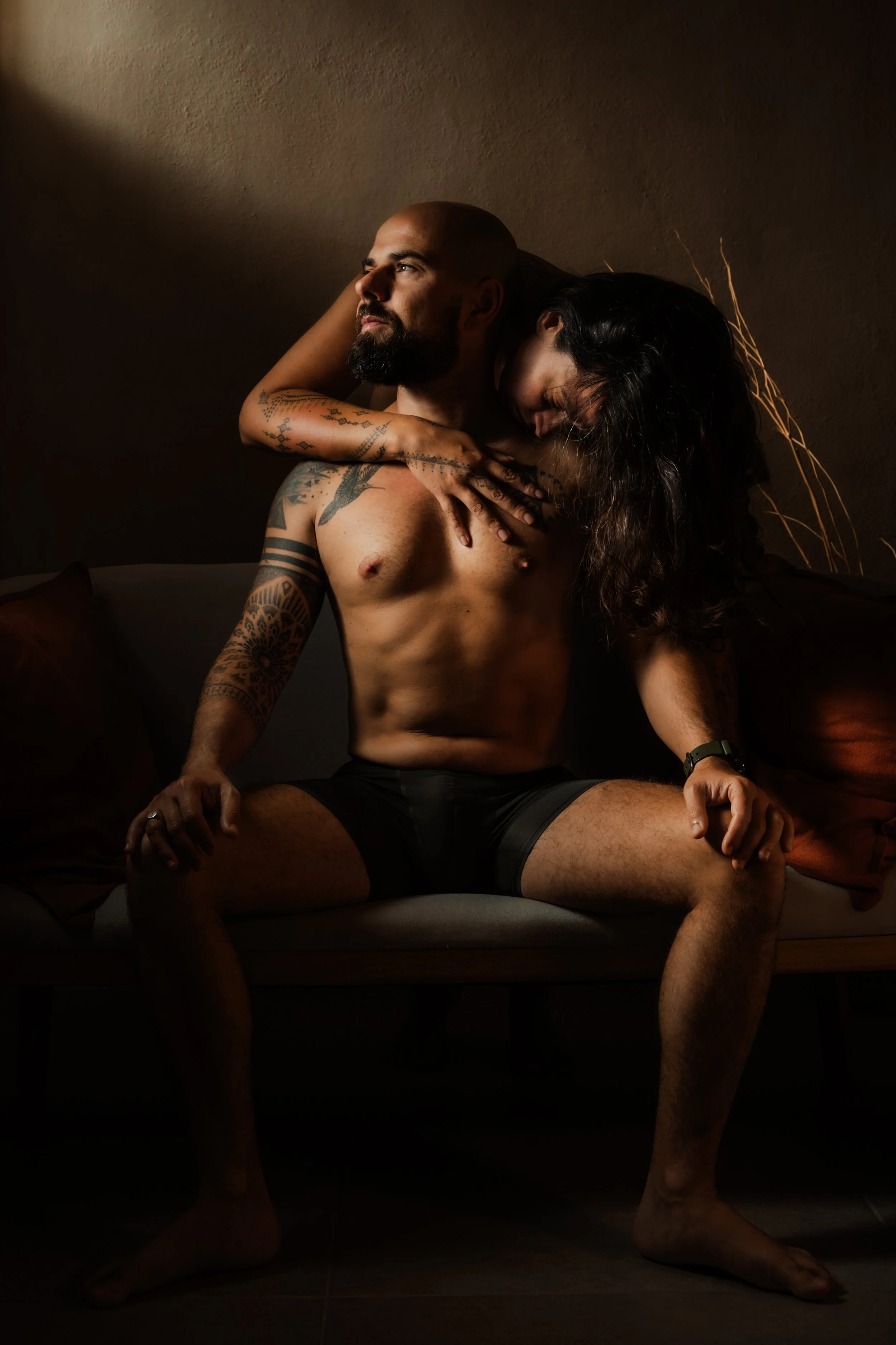 A shirtless man with tattoos sitting on a sofa with a woman embracing him from behind, her head resting on his shoulder in a dimly lit room.