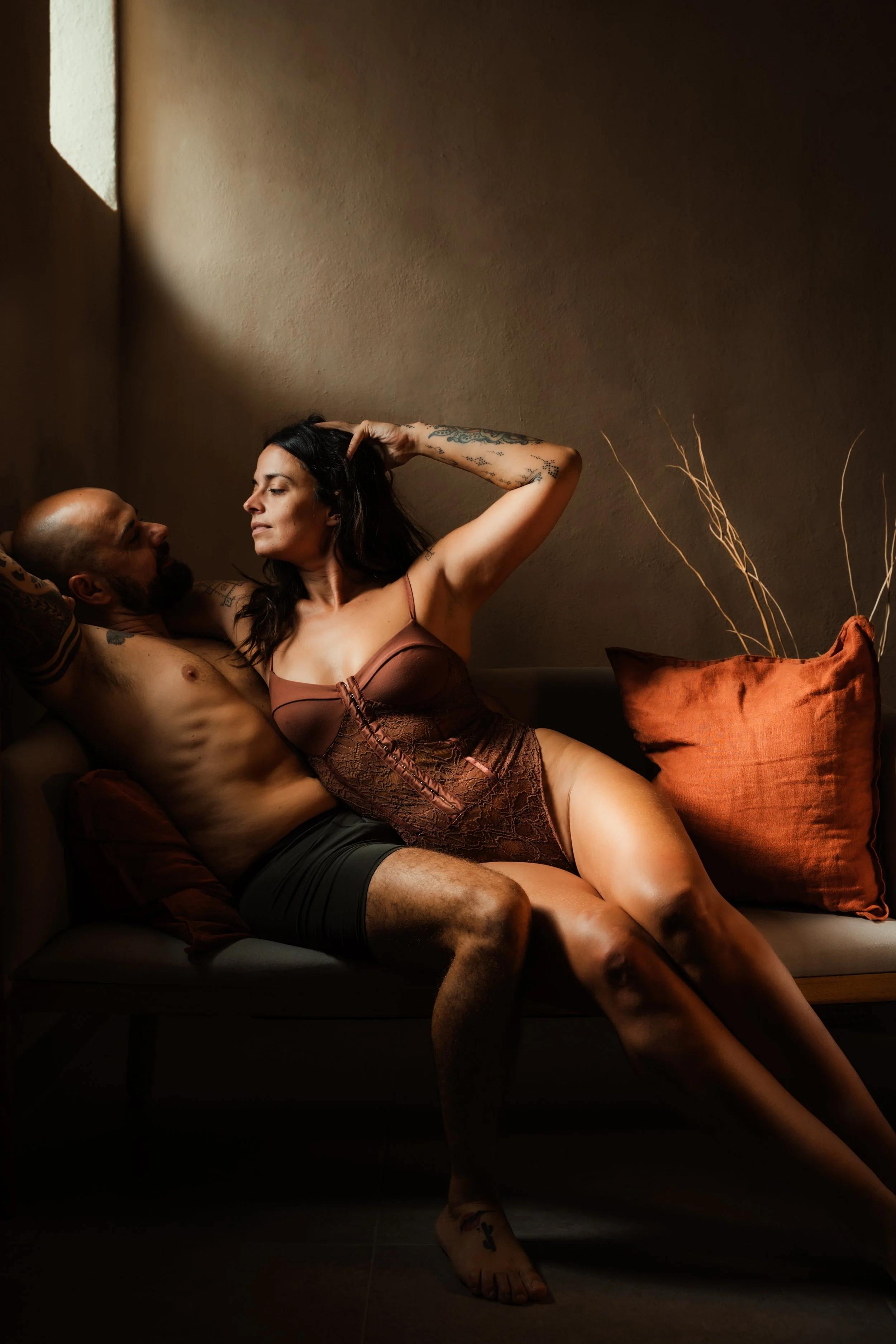 A man and woman in intimate pose on a sofa, with dim warm lighting and a plain wall background.