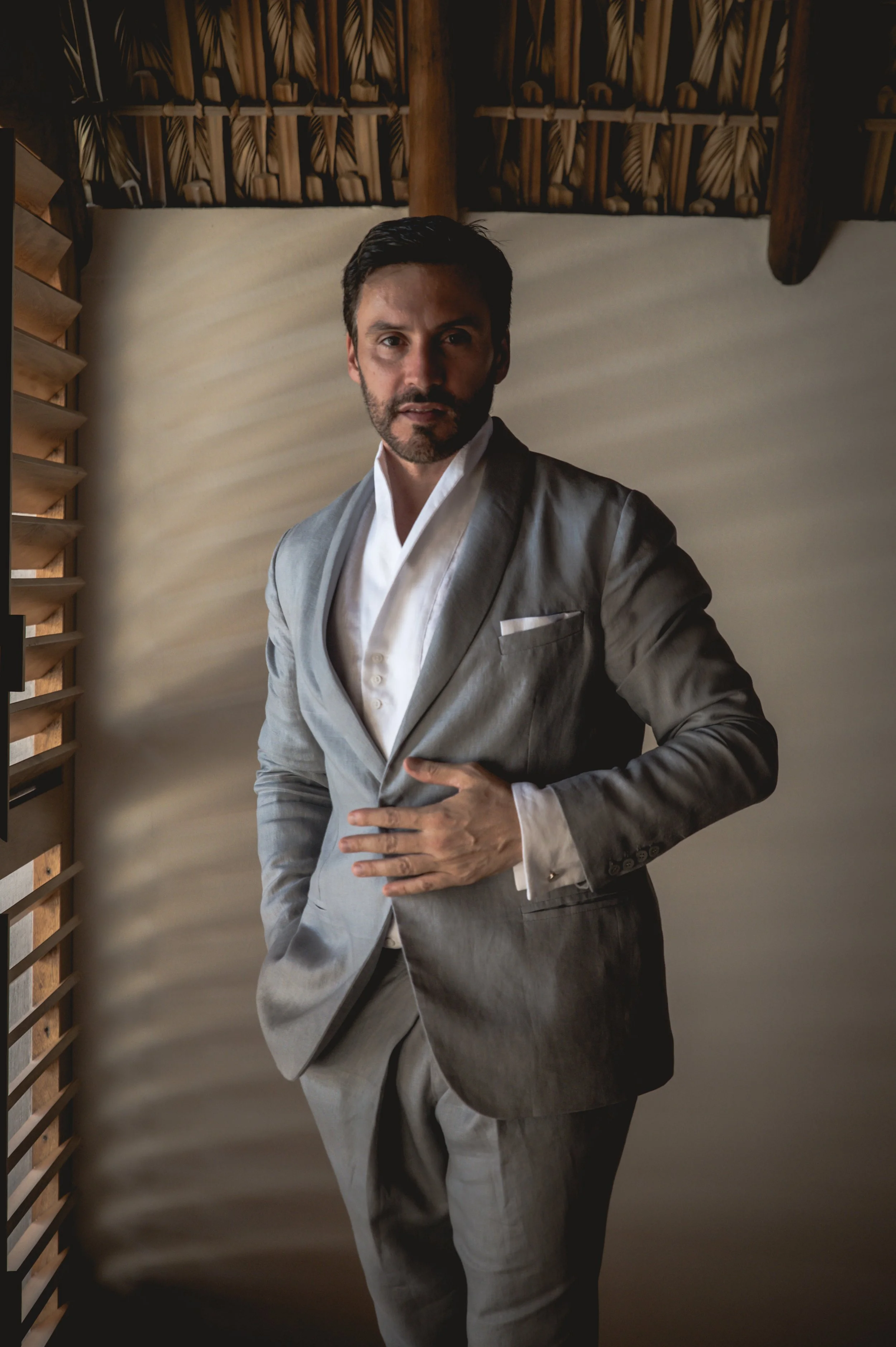 Beautiful profesional wedding photography of a Groom looking straight at the cemera with his suit on beside a window in a luxury villa in Puerto Escondido.