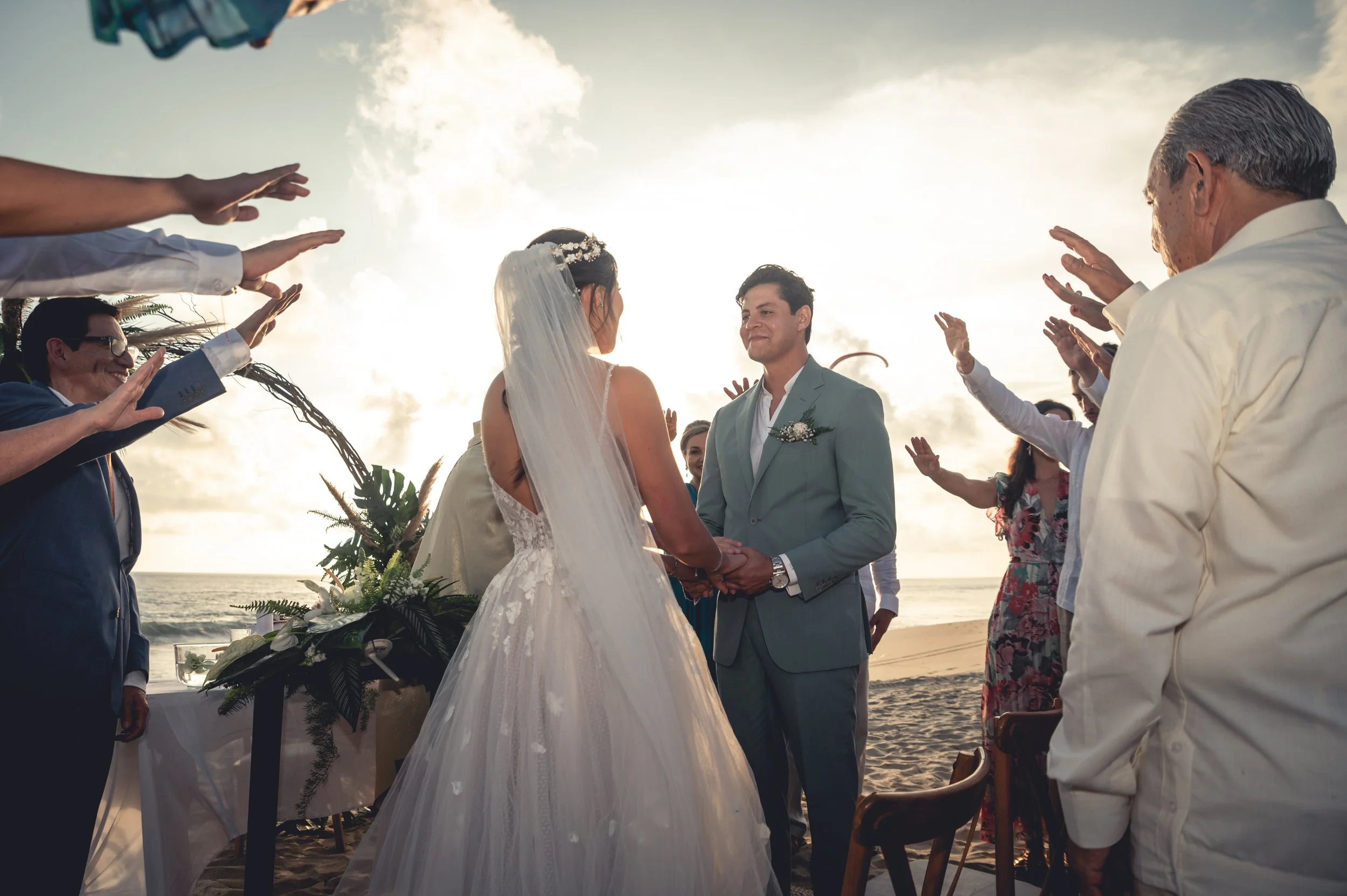 Wedding ceremony in Puerto Escondido where a Mexican couple from Monterrey is exchanging their vows in front a beautiful sunset at the beach in Puerto Escondido. Amazing frame with guest having their hands around the couple.