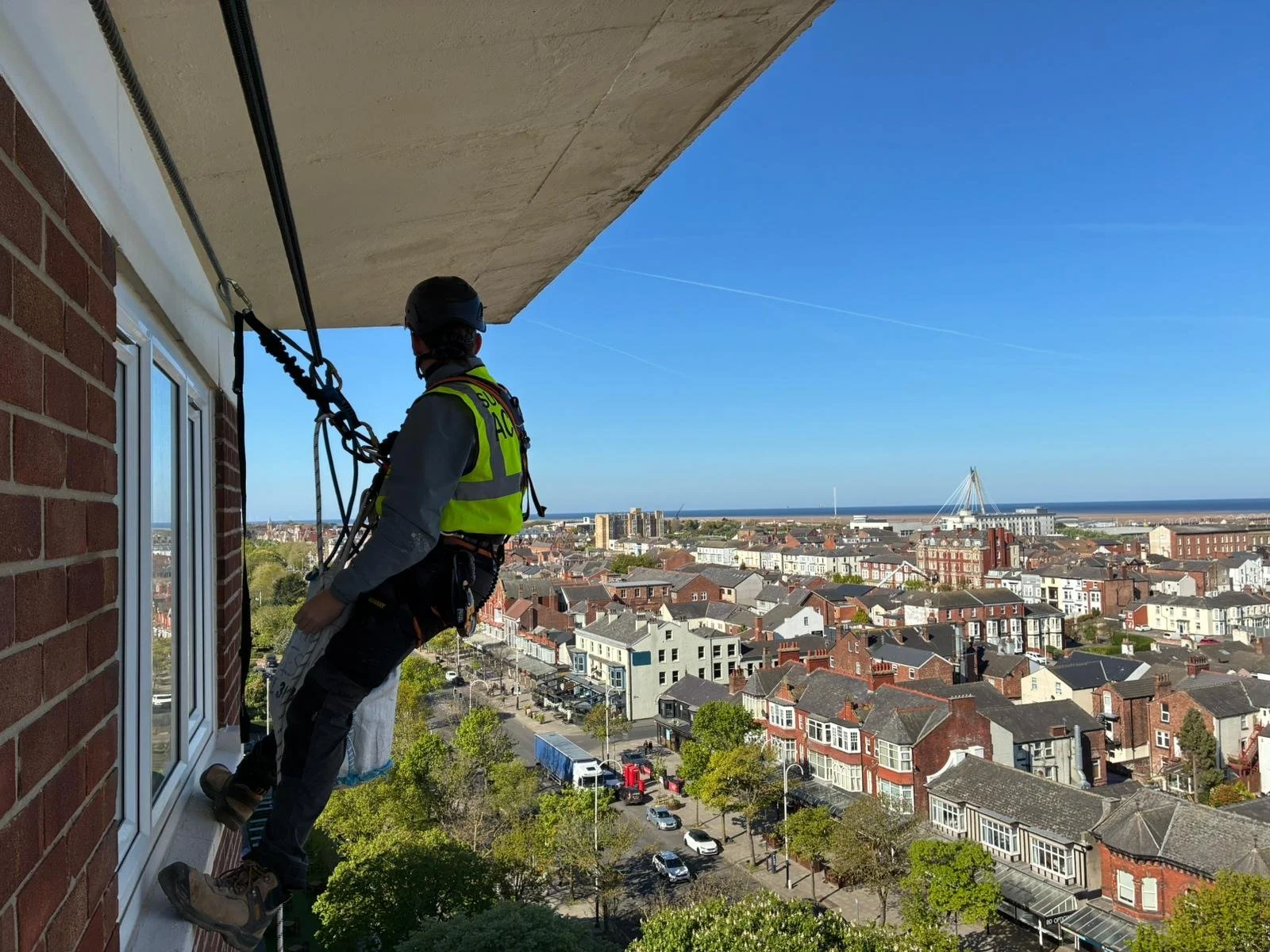A rope access technician accessing the side of a brick building. Further multi-storey buildings and a blue sky are seen in the background.