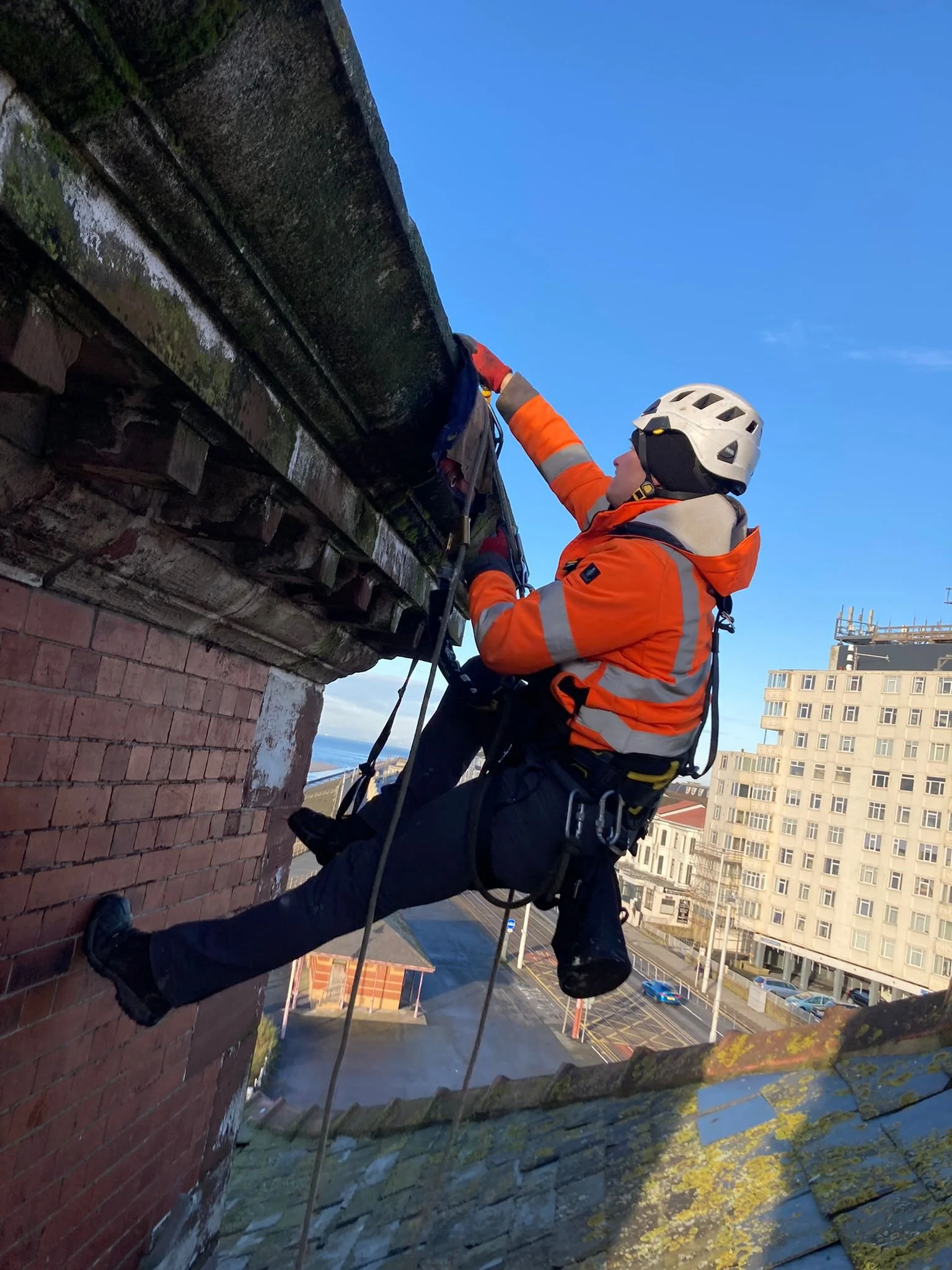 A rope access technician accessing the side of a brick building. Further multi-storey buildings and a blue sky are seen in the background.