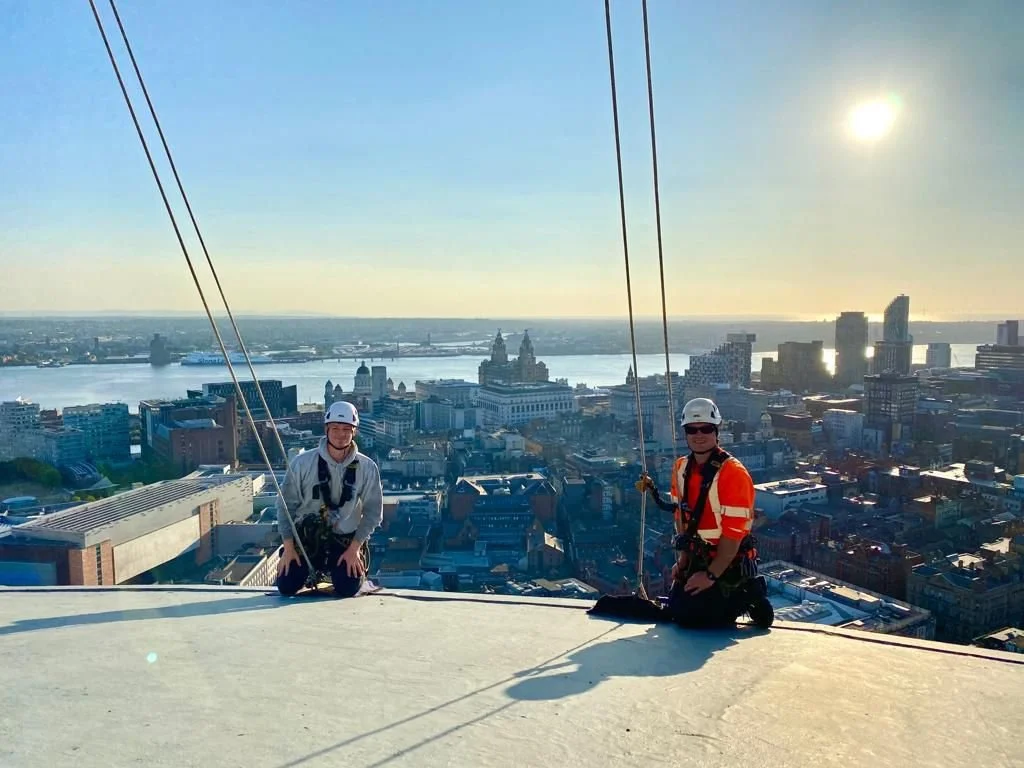 Two technicians are kneeling on a tall building roof, smiling. They are holding on to their ropes. The Liverpool skyline, the mersey, and the three graces buildings can be seen in the background.