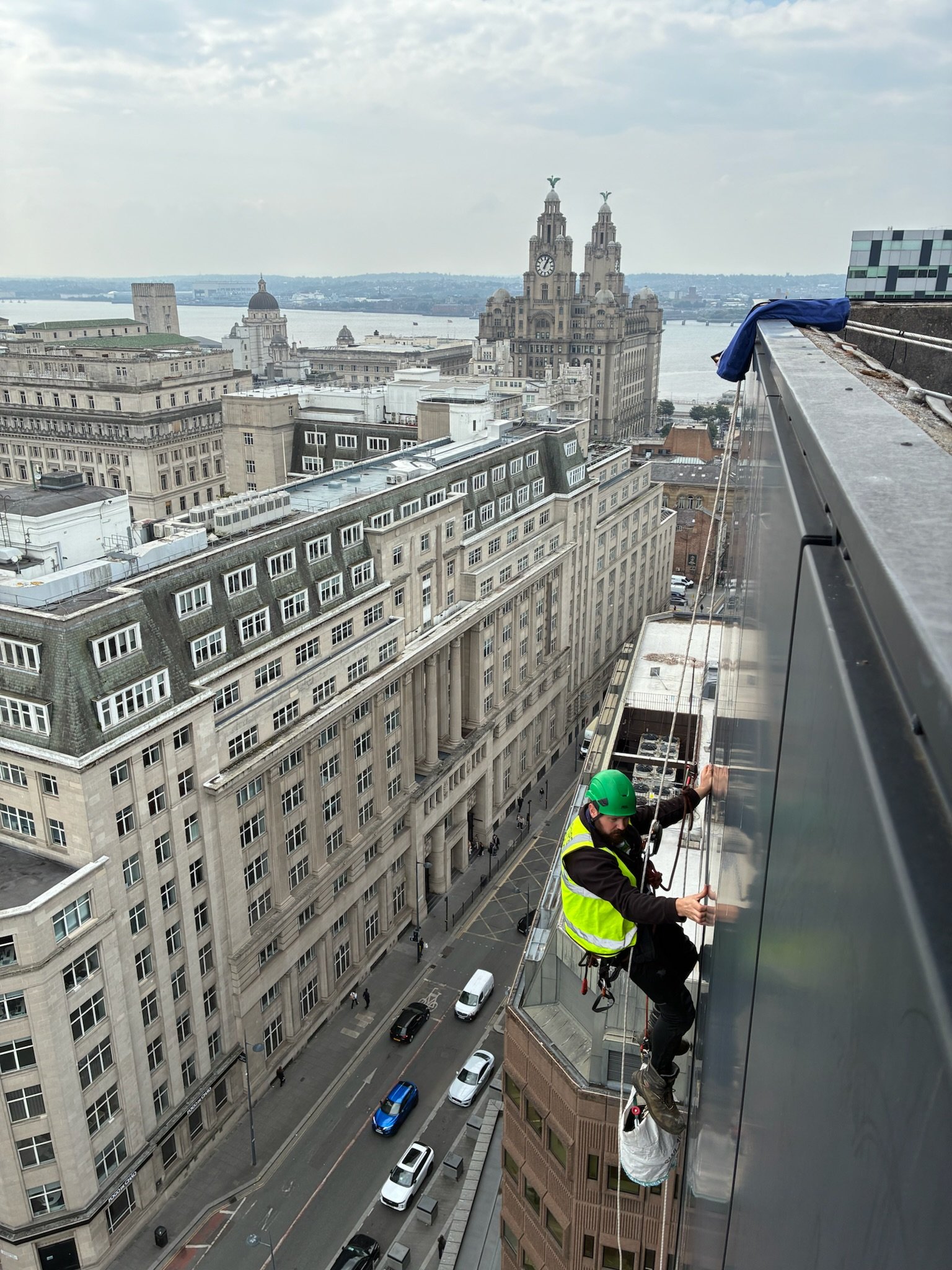 A birds-eye view of a rope access technician working on a large green metal bridge. A black tarmac road with white markings can be seen running beneath the Bridge.