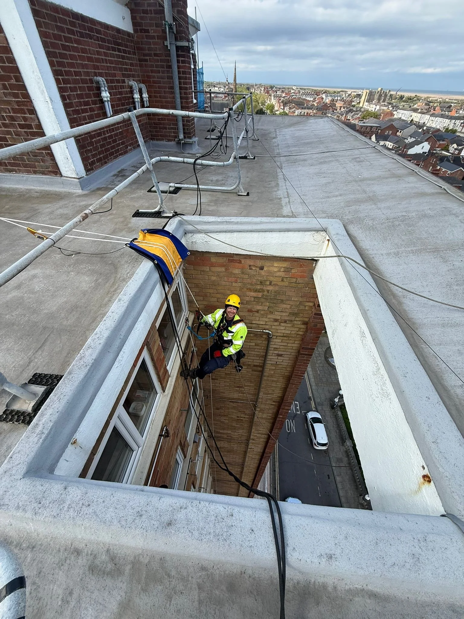 Two technicians are kneeling on a tall building roof, smiling. They are holding on to their ropes. The Liverpool skyline, the mersey, and the three graces buildings can be seen in the background.