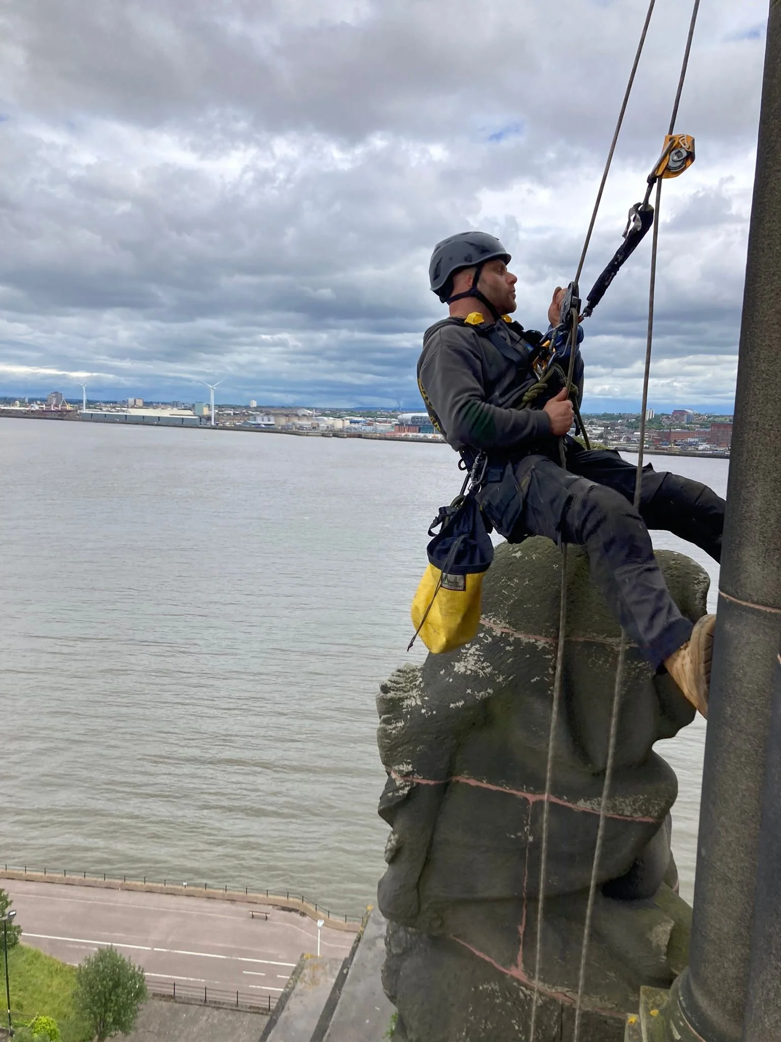 A rope access technician with a safety helmets sits on a large tower or stone or rock. A river can be seen in the background and he appears to be working on or repairing a large metal pillar.