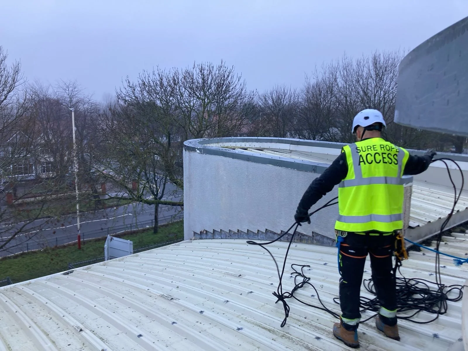 A rope access technician works on the roof of KGV college. He wears a 'Sure Rope Access' hi viz vest, protective gear, and a helmet.