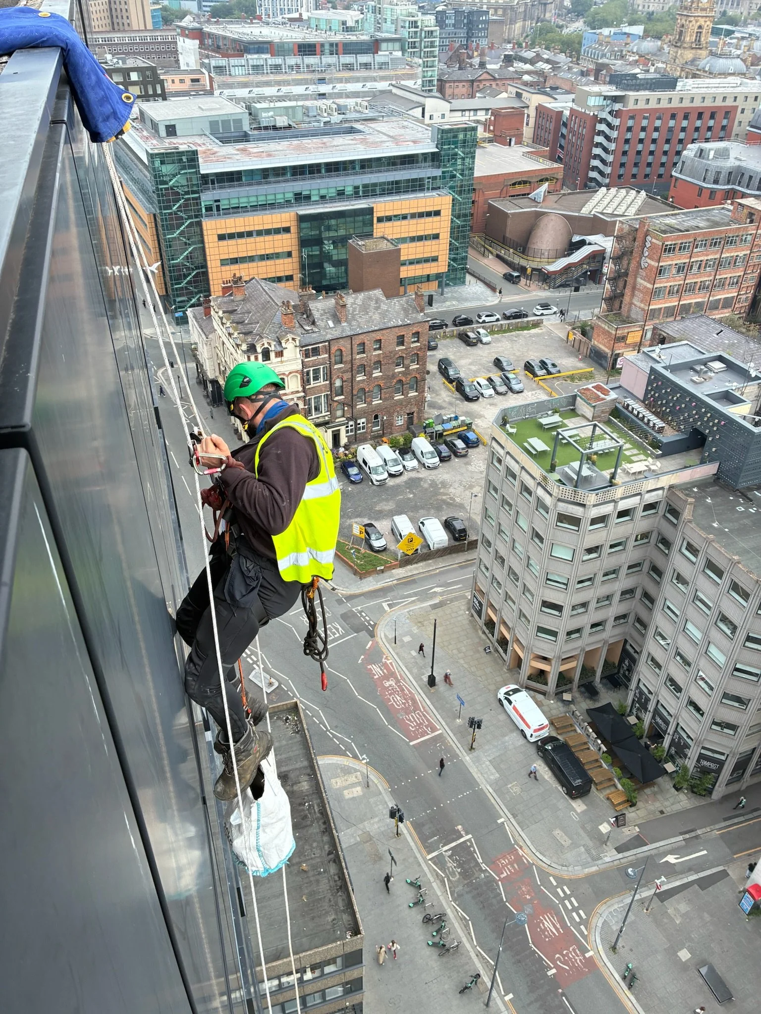 A worker wearing safety gear and a helmet is sitting on a green metal bridge, smiling.