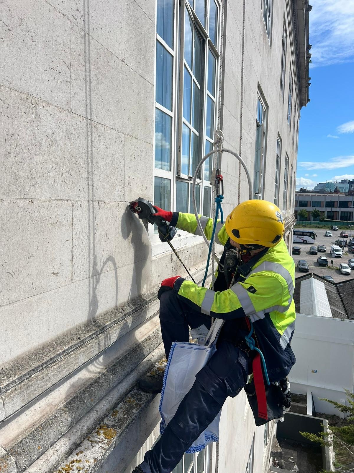A rope access technician with a safety helmets sits on a large tower or stone or rock. A river can be seen in the background and he appears to be working on or repairing a large metal pillar.