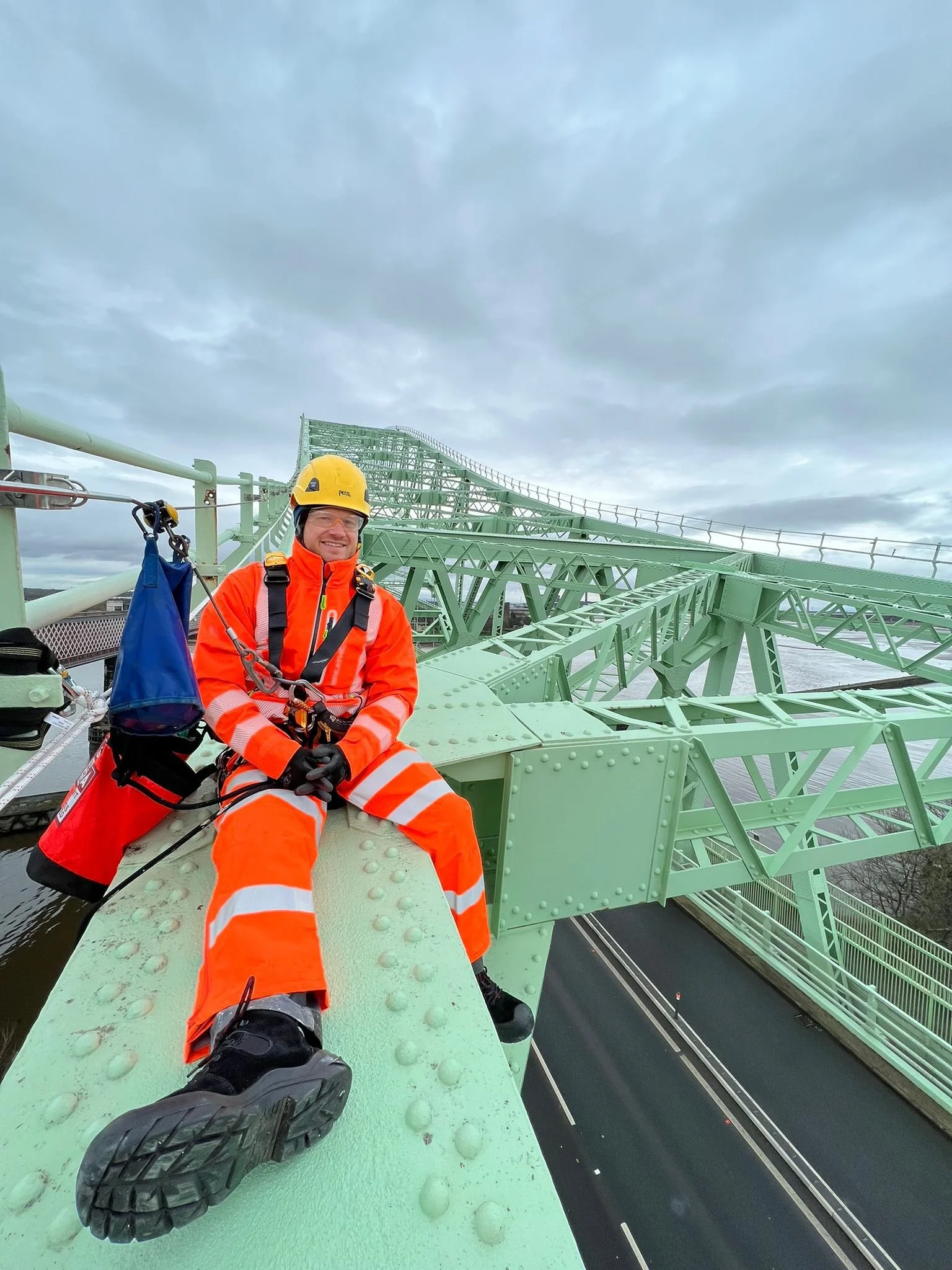 A worker wearing safety gear and a helmet is sitting on a green metal bridge, smiling.