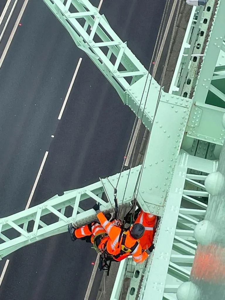 A birds-eye view of a rope access technician working on a large green metal bridge. A black tarmac road with white markings can be seen running beneath the Bridge.
