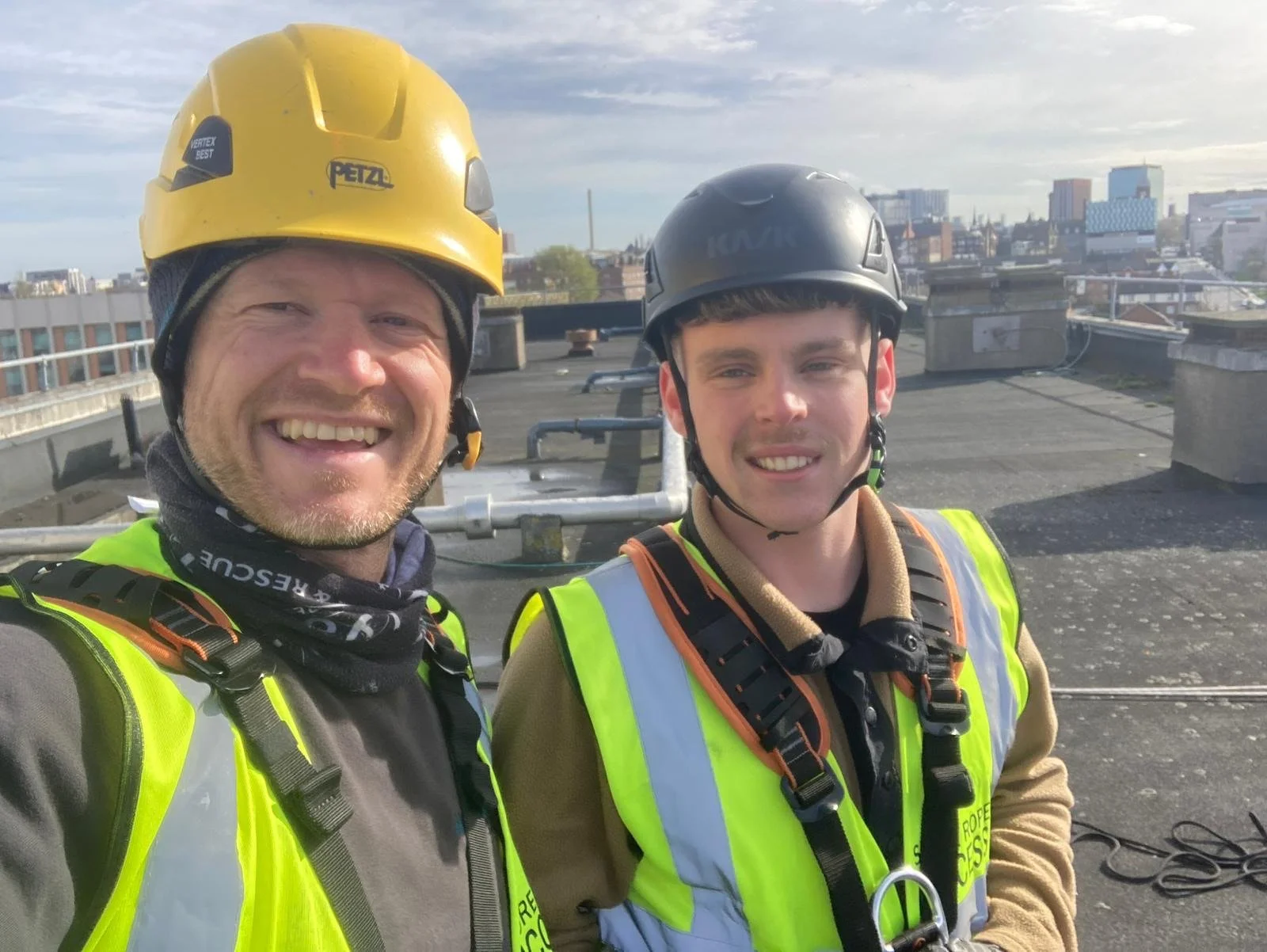 Two rope access technicians are kneeling on the roof of a tall building, smiling at the camera. Liverpool city skyline and the river mersey are in the background. The sun is shining. 