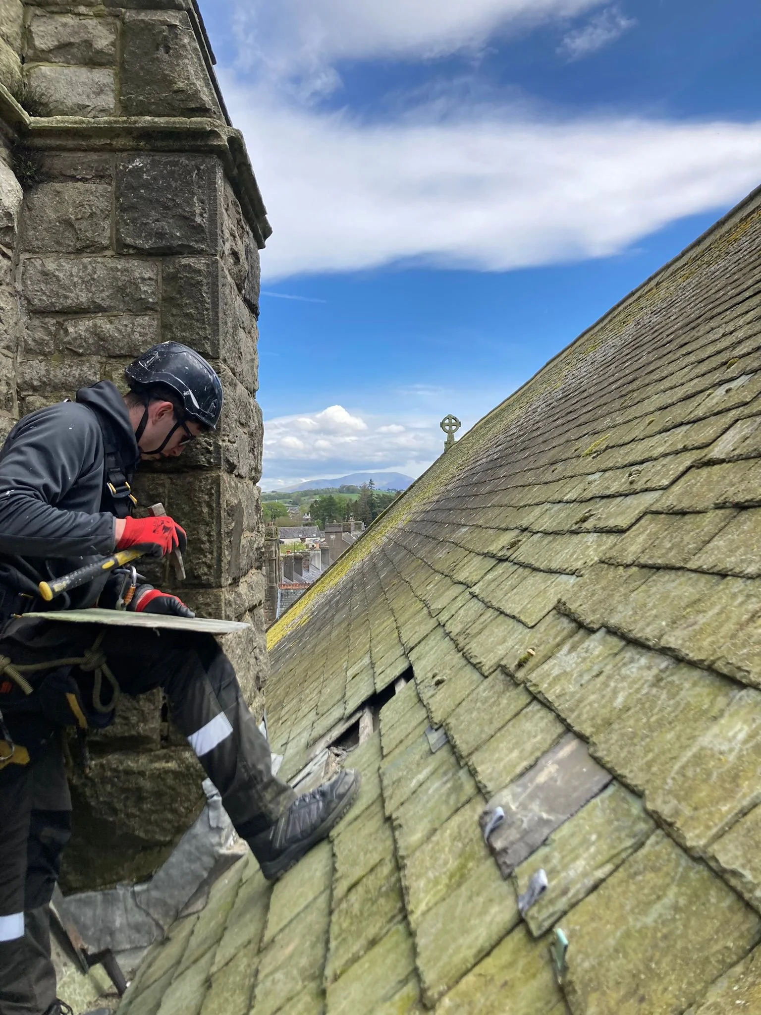 A technician wearing a helmets sits on a tall stone church roof, with a chimney or tower behind him. He is repairing a piece of slate.