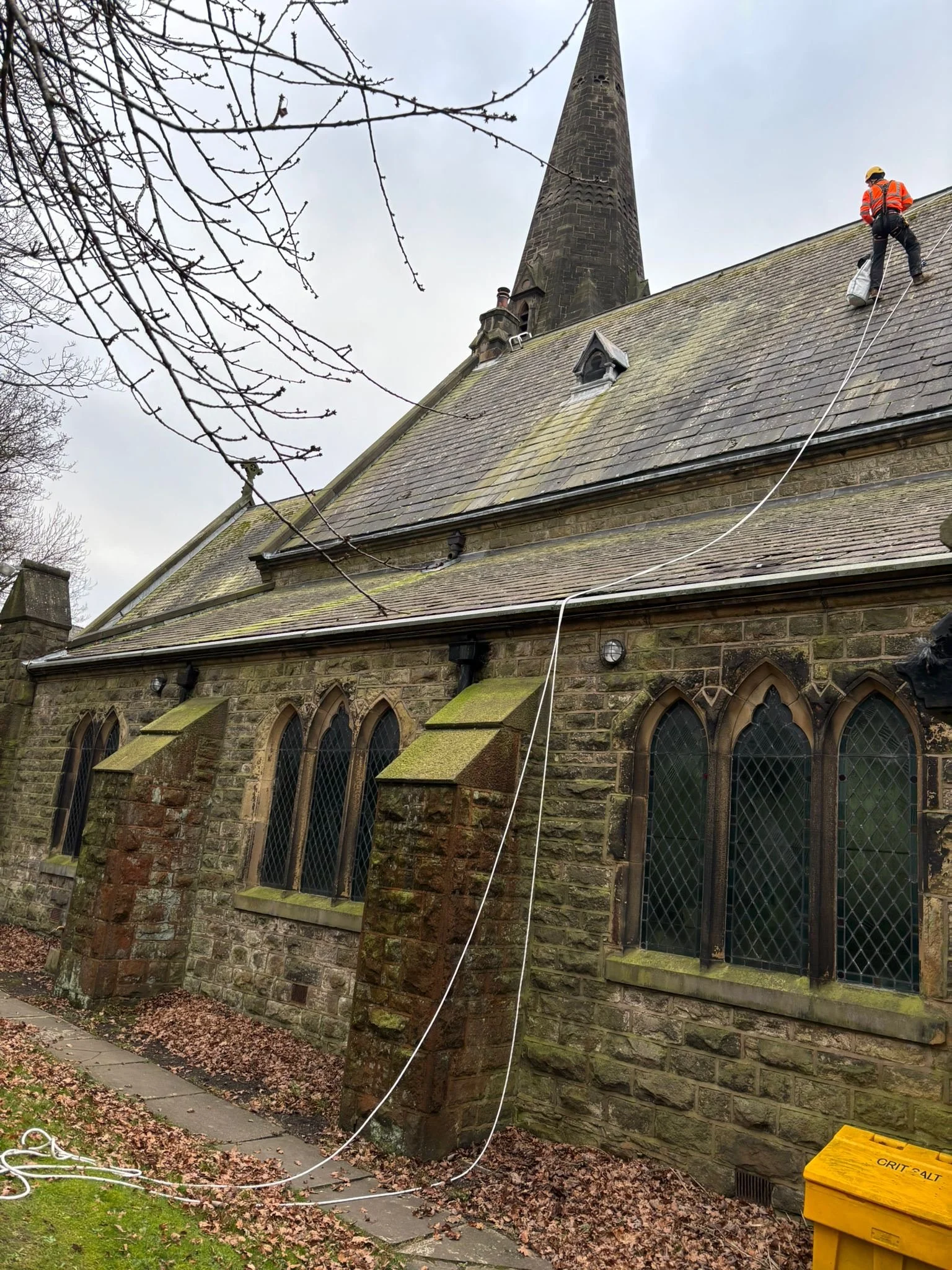 A technician wearing a helmets sits on a tall stone church roof, with a chimney or tower behind him. He is repairing a piece of slate.