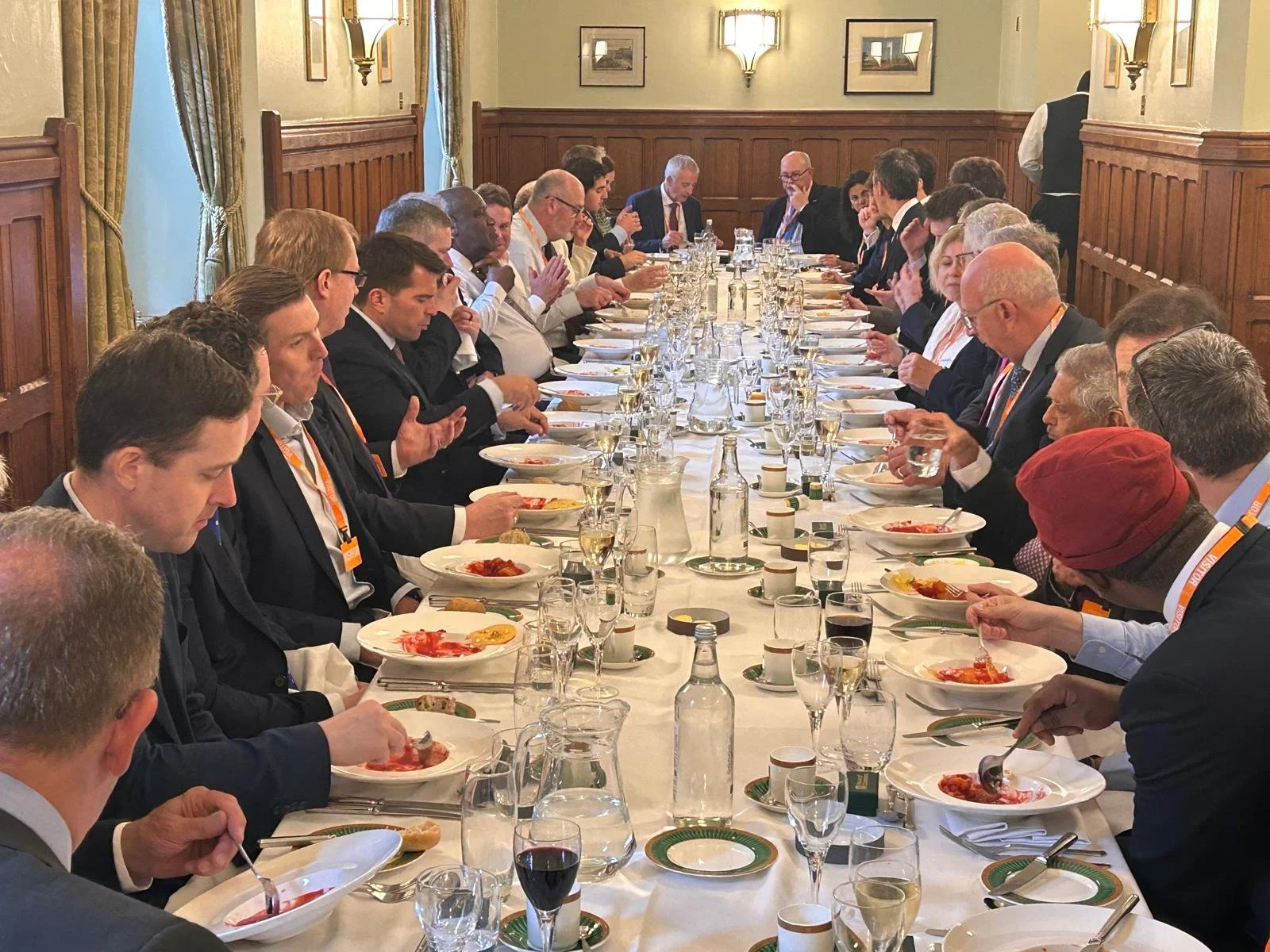 A large group of people seated at a long dining table in a formal setting, eating a meal, with various glasses, plates, and bottles on the table.