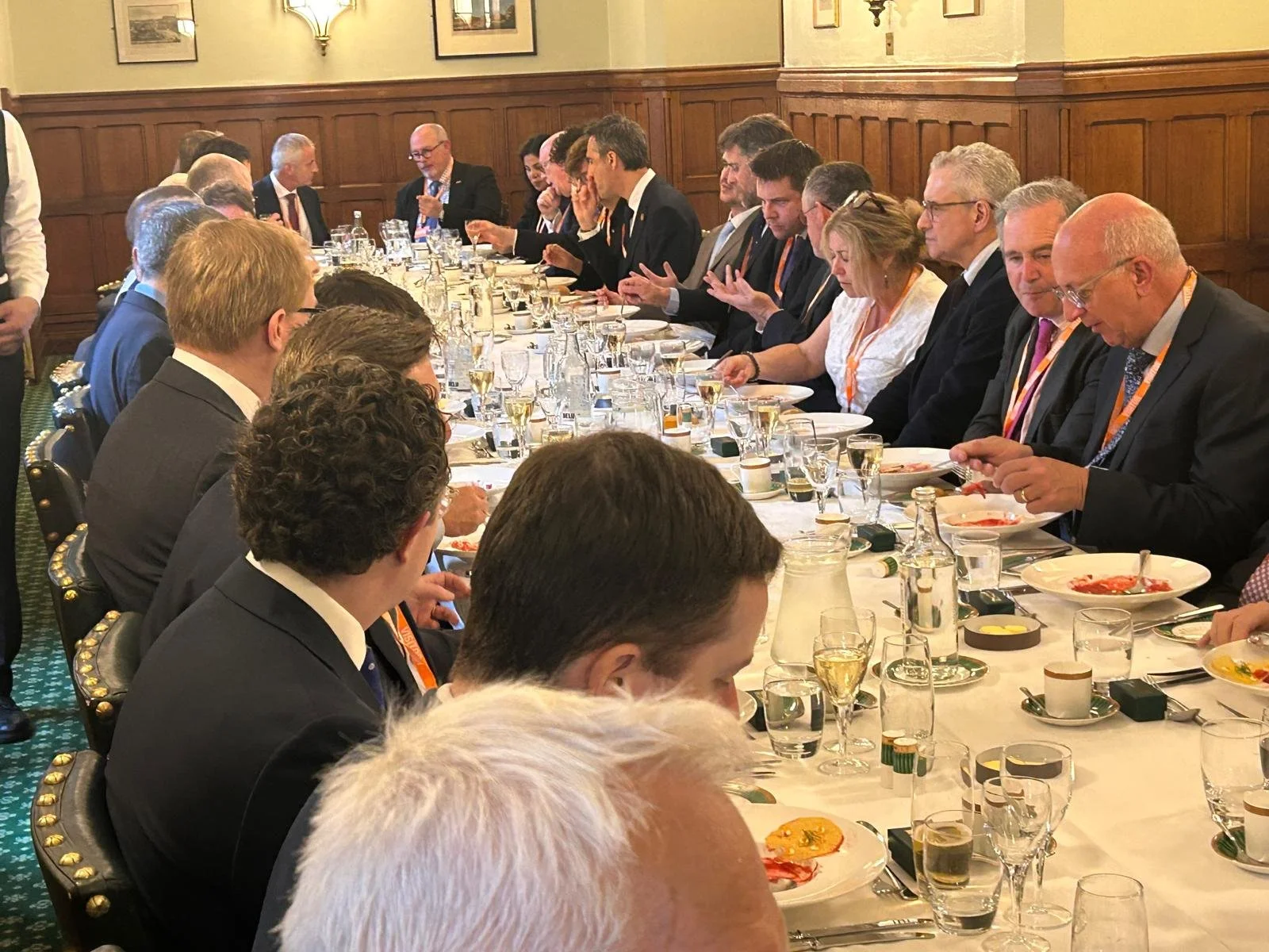 A formal dinner with many people seated at a long table in a wood-paneled room, engaged in conversation. The table is set with glasses, plates, and silverware, and some people are eating while others are talking.