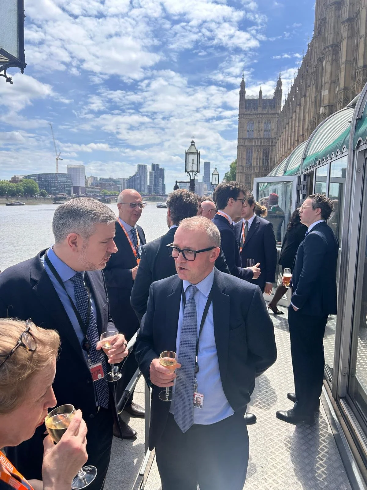 Business professionals in suits and glasses socializing with drinks on a boat deck near the River Thames, with the Palace of Westminster and modern buildings in the background on a partly cloudy day.