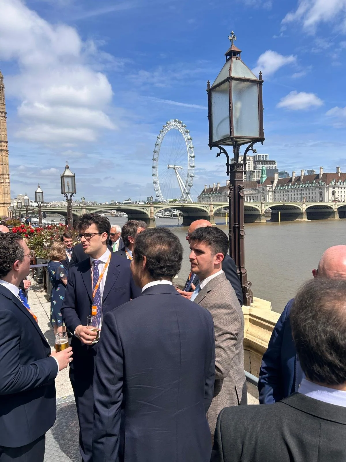 A group of people dressed in business attire, standing and talking outdoors near the River Thames in London. The London Eye Ferris wheel is visible in the background against a blue sky with some clouds, along with the iconic London skyline.