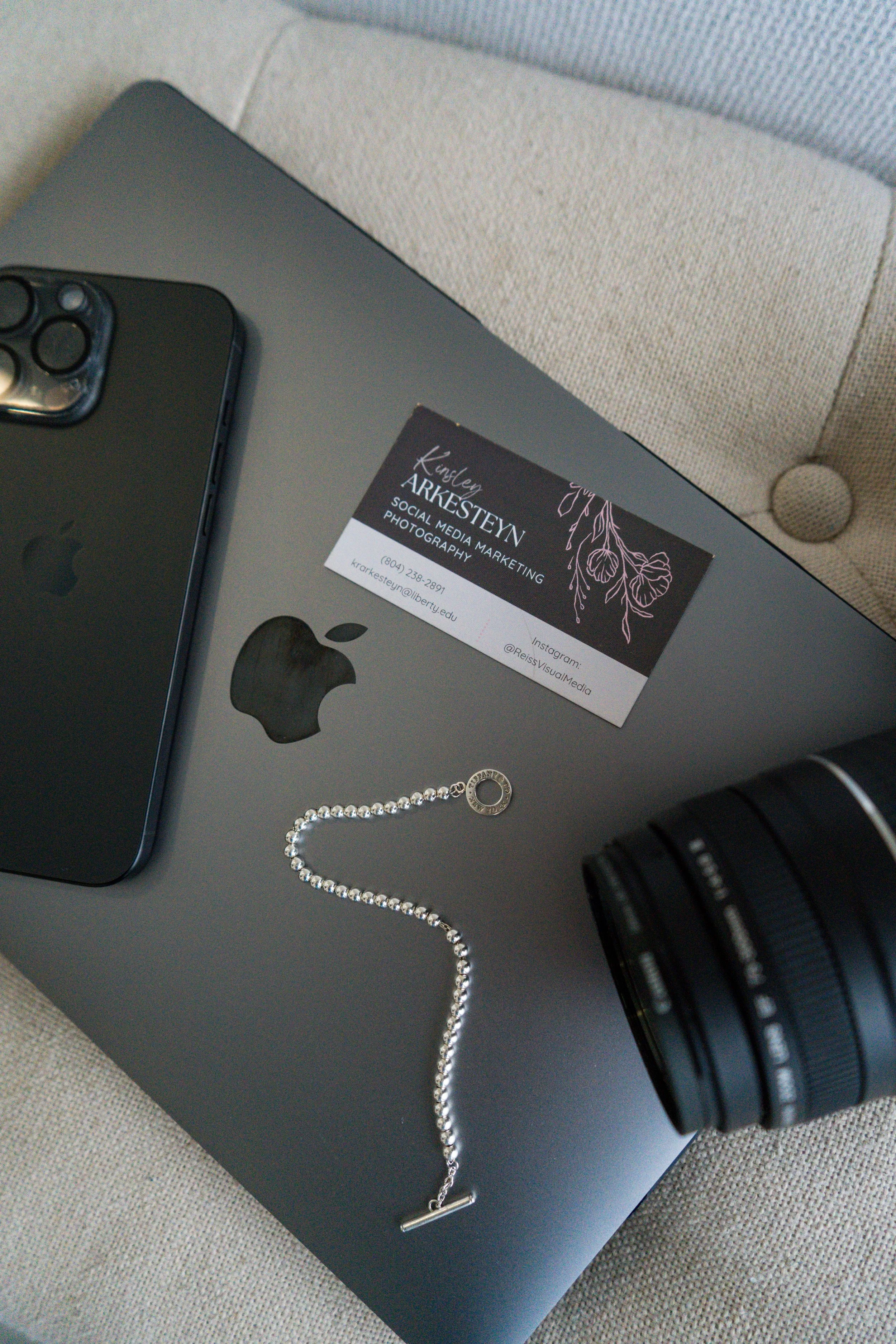 Top-down view of a MacBook with a business card, an iPhone, a camera lens, and a silver jewelry chain on the laptop surface.