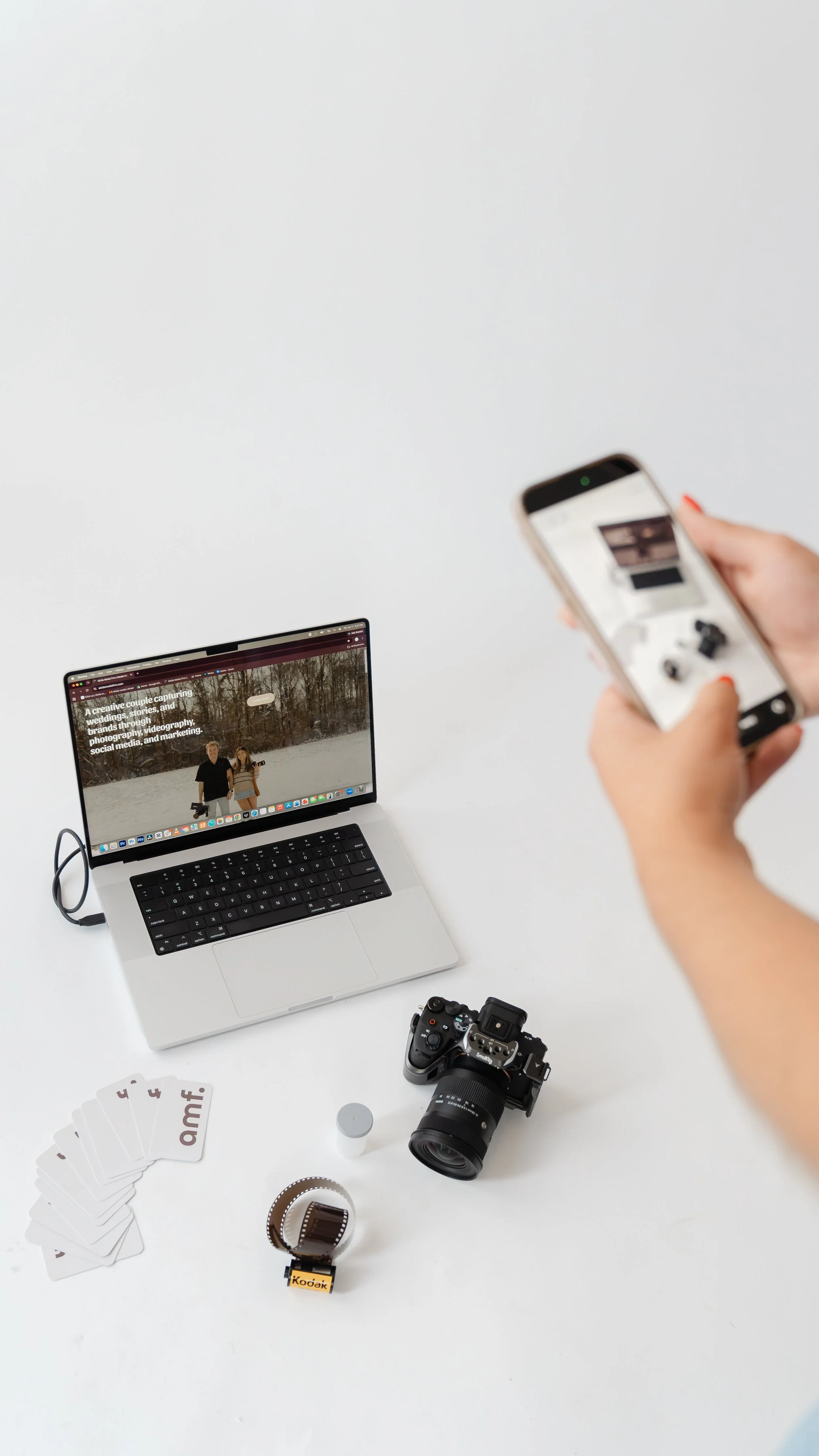 Top-down view of a MacBook with a business card, an iPhone, a camera lens, and a silver jewelry chain on the laptop surface.