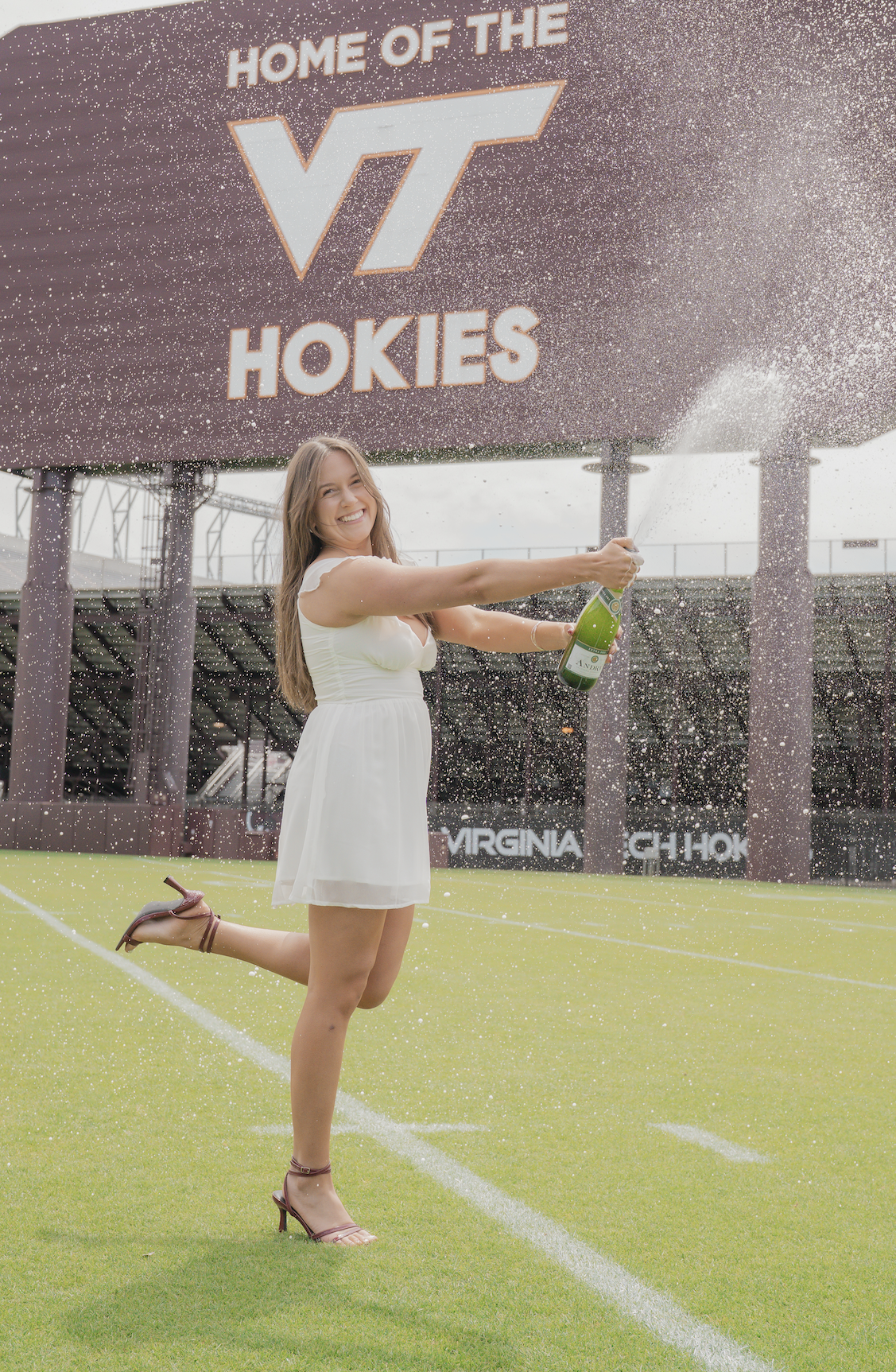 A young woman in a white dress and red heels sitting on a large red LOVE sign outside a brick building during daytime, holding a graduation cap.