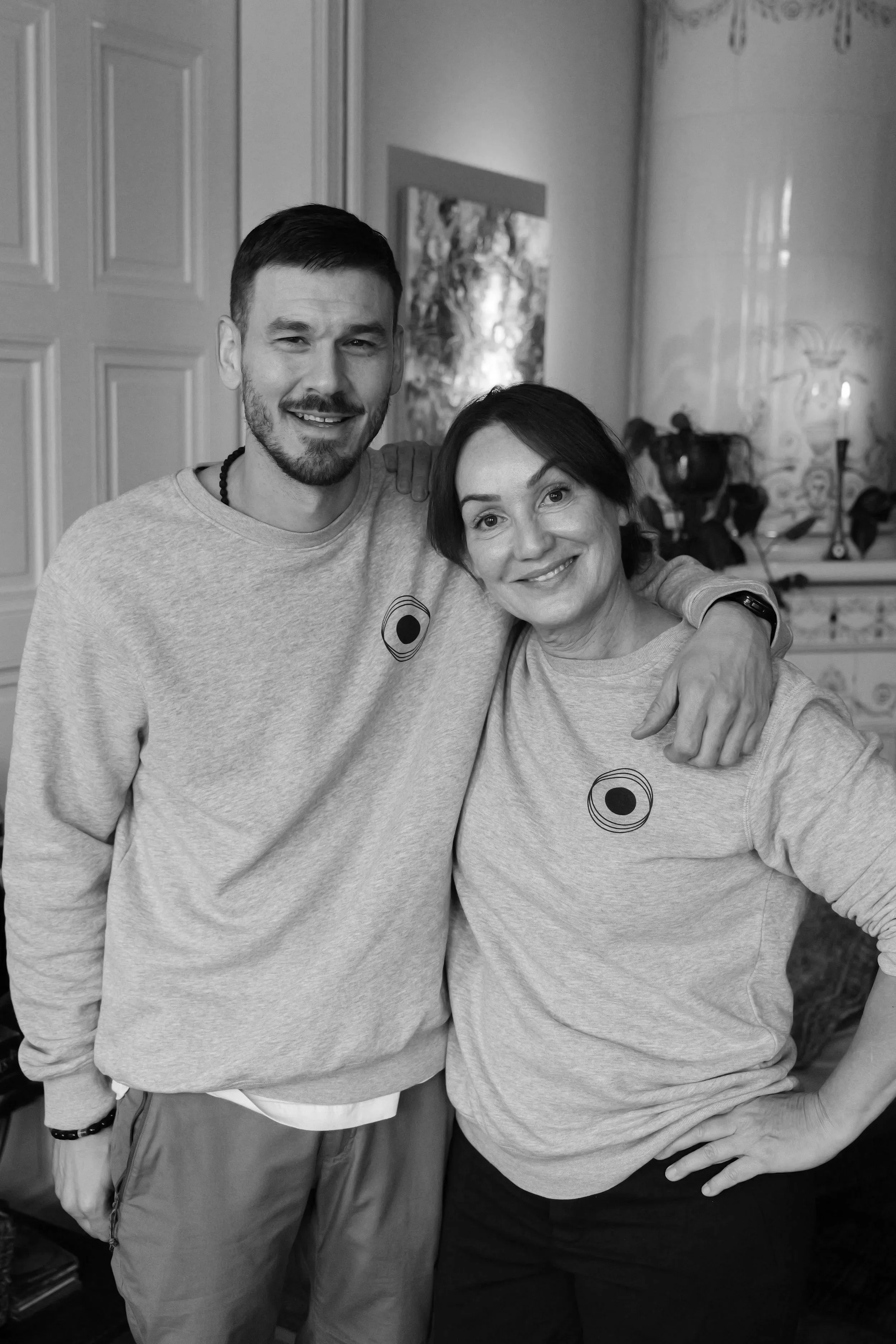 Två personer i grå tröjor med cirkelmönster, en man och en kvinna, som står nära varandra och ler mot kameran, i ett hemmet.