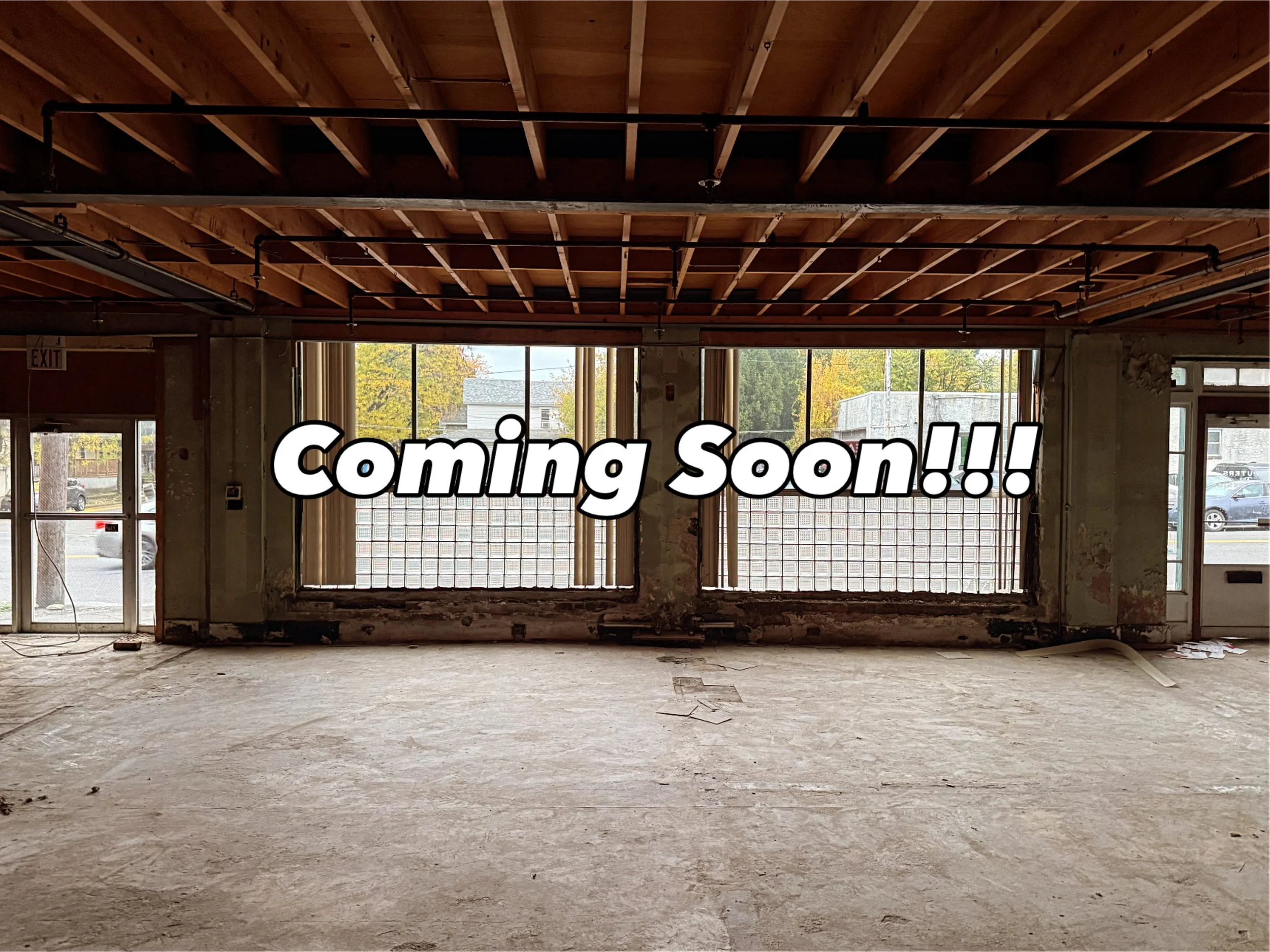 Empty storefront space with large front windows, exposed ceiling with wooden beams, and concrete floor, with the text 'Coming Soon!!!' overlayed.