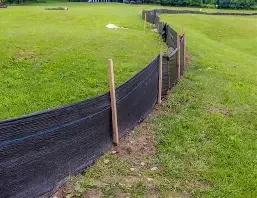 A black plastic fence supported by wooden stakes, running along a grassy area with soccer field markings in the background.