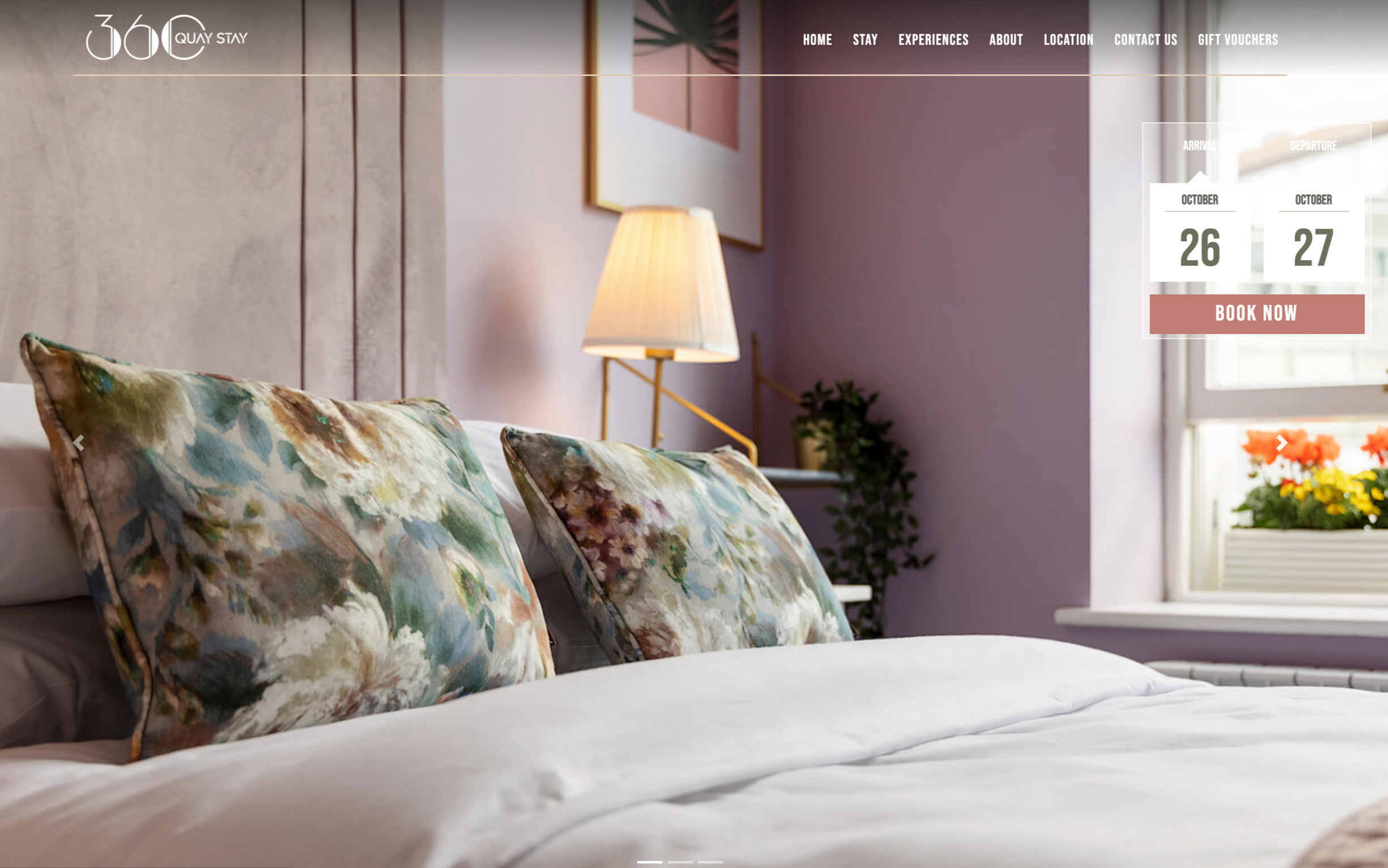 A floral cushion lying on a large bed, next to a flower-laded windowsill in a lavender themed room.