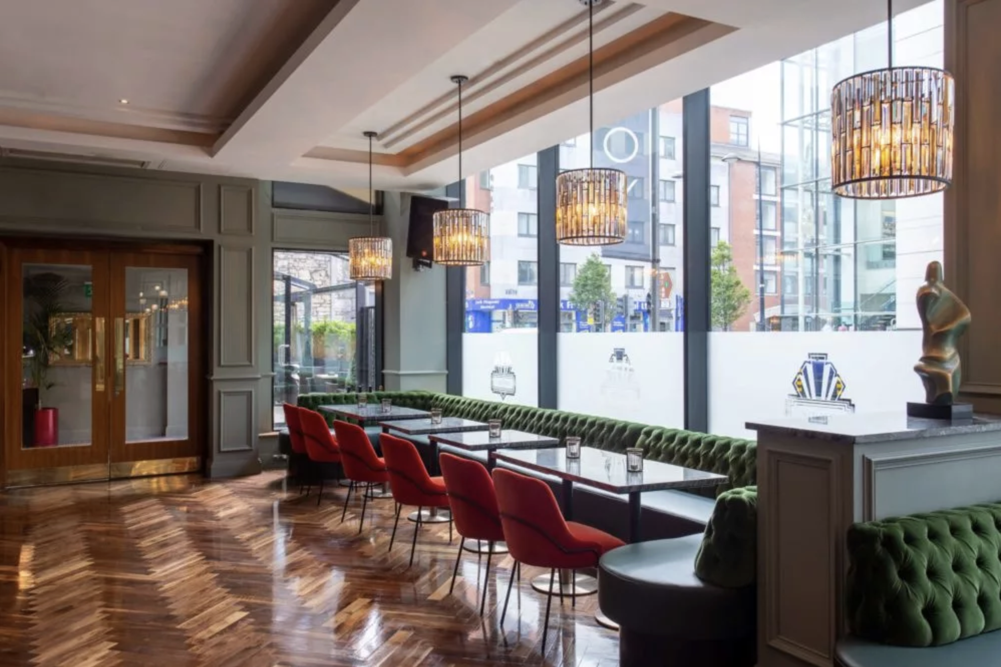 Corner of the restaurant with five red chairs, five tables, a green leather bench and large windows.