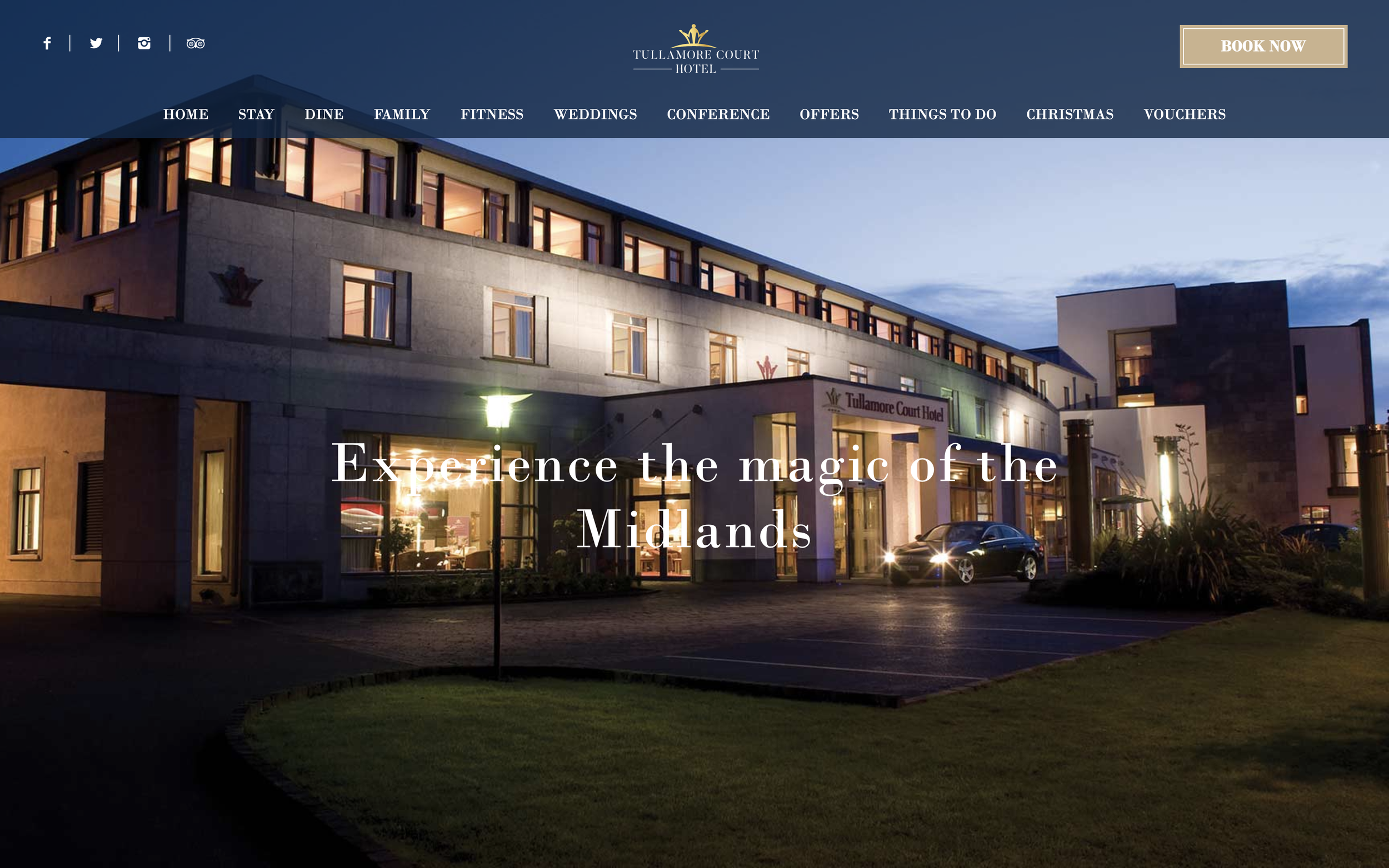 Exterior of the front of a three storey hotel, lit up at night with a luxury black car out front.