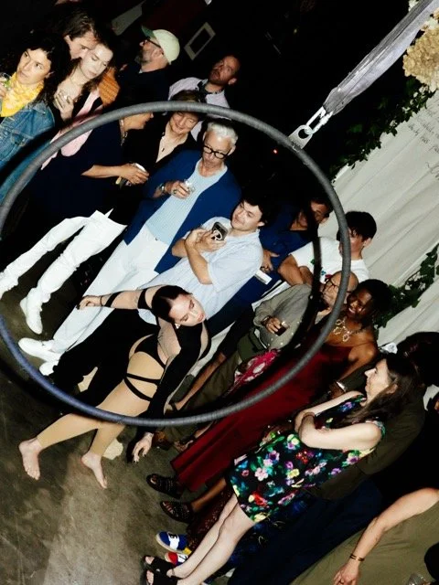 Group of people gathered at a party, some are taking pictures and others are talking, with white curtains and floral decorations in the background.