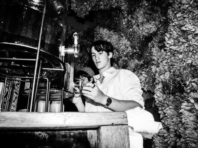 A young man with dark hair pouring a beverage from a tap at a rustic outdoor bar, with a background of dense foliage.
