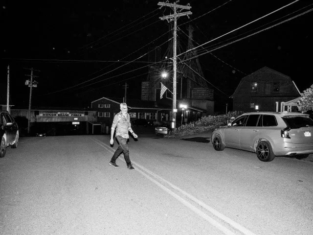A man crossing a street at night in a small town with parked cars and buildings in the background.