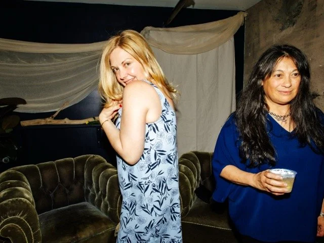 Two women at a social gathering, one with blonde hair in a light blue dress with a floral pattern, and the other with dark hair in a blue shirt holding a drink, standing in a dimly lit room with a velvet couch and decorative fabric drapes.