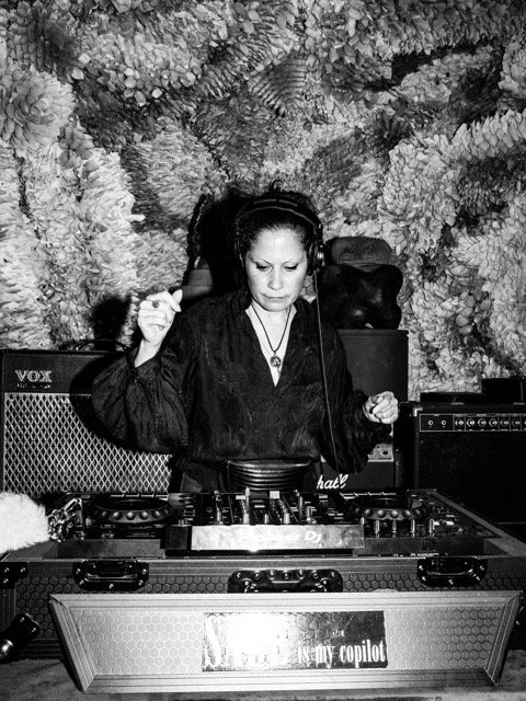 A female DJ with headphones at a DJ booth in front of a textured rock wall, mixing music.