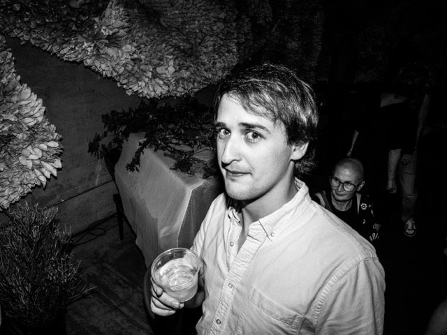 A young man with light skin and dark hair holding a drink in his right hand, looking at the camera, at a social gathering or party with floral decorations and other people in the background, in black and white.