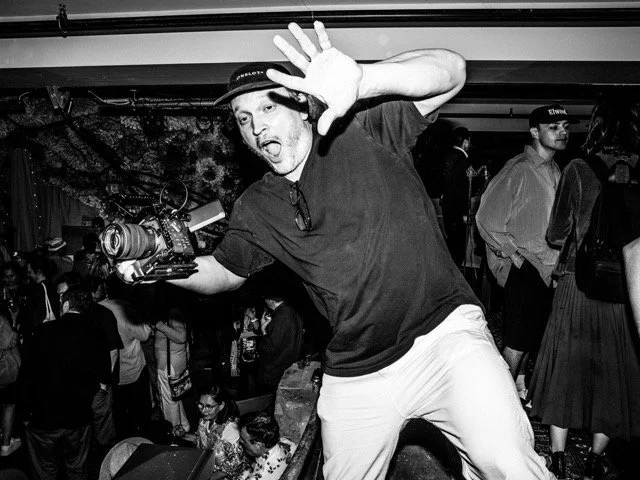 A man with a camera on a stabilizer, wearing headphones, raising one hand and making a playful face at a crowded party or event.