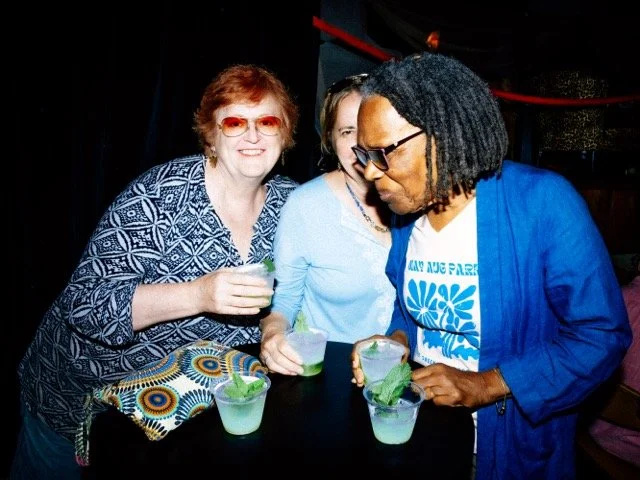 Three women at a bar holding drinks, smiling and chatting.