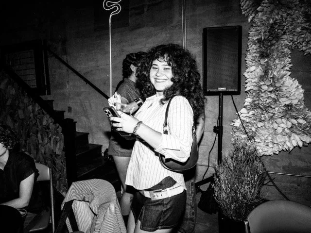 A young woman with curly hair smiling and looking at her phone in a social setting with other people and decorative plants in the background.