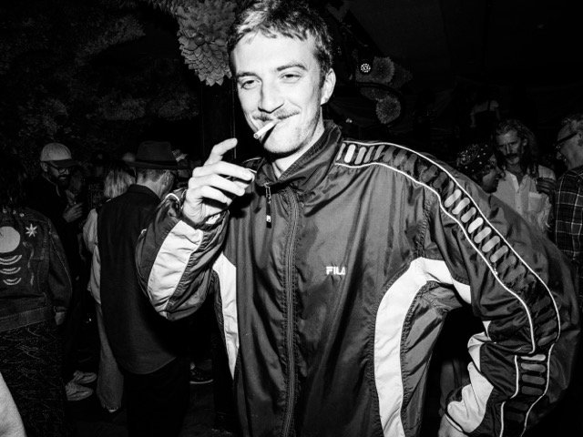 A young man smiling and holding a cigarette in his right hand at a social gathering, wearing a FILA sports jacket with other people in the background.