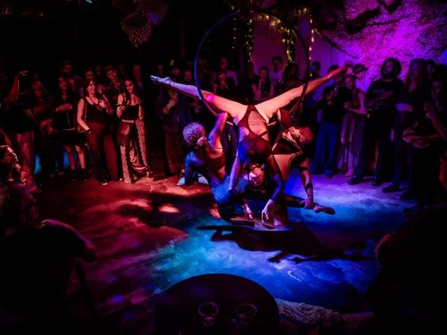 Performers doing an acrobatic dance under colorful lights at a nightclub or dance event, with an audience watching.